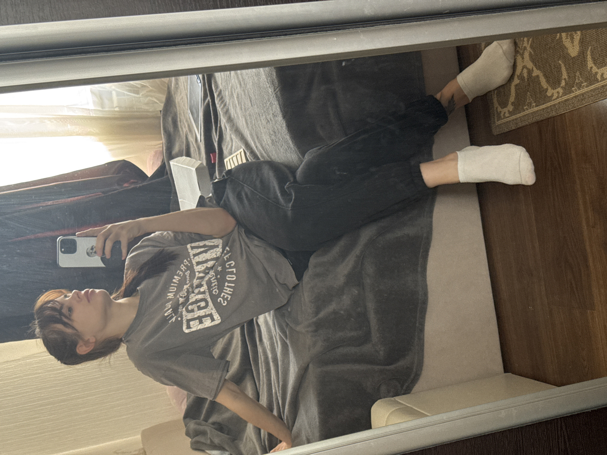 A person wearing a gray t-shirt and dark pants is lying on a gray blanket with white socks, reflecting off of a mirror. The area appears to be a bedroom, as a bed is visible. There is also a portion of a rug on the floor and a wooden floor. The location in the city of Rostov-on-Don, Russia, cannot be verified as it is unknown.