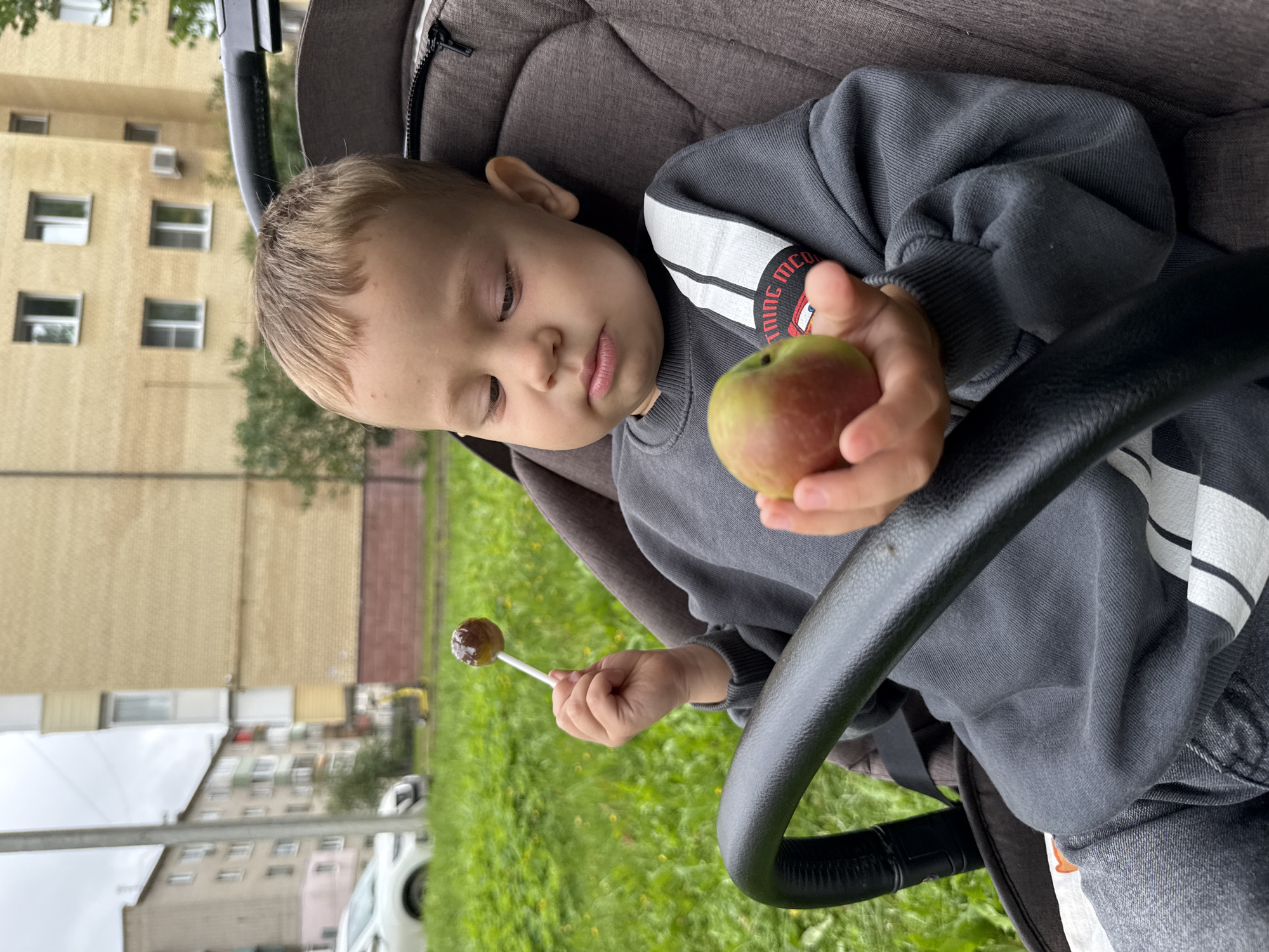 A young Caucasian child, appearing to be between two and three years old, is seated in a brown stroller outdoors. The child has light brown hair and is wearing a gray sweatshirt. In their right hand, the child holds a small, round piece of fruit, possibly an apple, which is green and red. Their left hand holds a lollipop on a white stick. The child is looking down at the fruit.
The background suggests an urban environment, likely in Rusanovo, Russia. To the left, a white car is parked on a street, and behind it are multi-story apartment buildings with visible windows. Green grass, with some small yellow flowers, covers the ground. A tall, thin pole, possibly a lamppost or utility pole, is visible on the left side of the frame. The sky appears overcast. The stroller's frame, a black handle, is in the foreground.