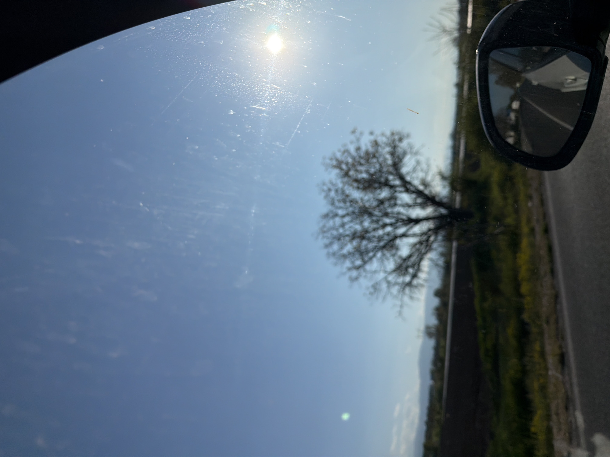 The image shows a view through a car window on a sunny day. The sky is mostly clear blue, with the sun visible. There is a tree with bare branches in the midground. A side mirror of the car is visible in the upper right corner, reflecting the road and roadside vegetation. The location is Unknown, Unknown.