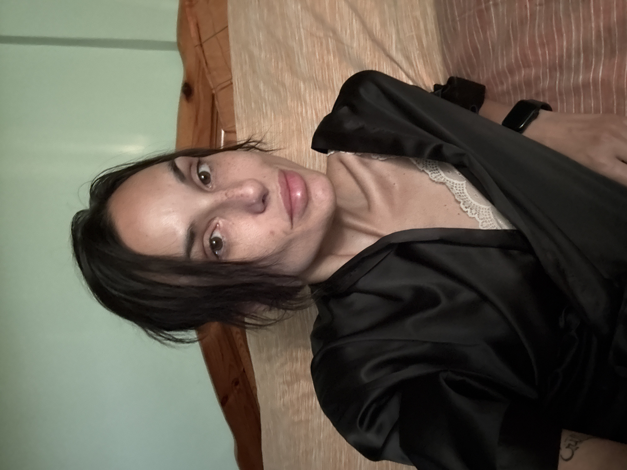 A close-up, frontal shot shows a woman with dark, shoulder-length hair, fair skin with freckles, and brown eyes. She is wearing a black satin robe over a white lace bralette. Her lips are slightly parted, and she is looking directly at the camera. The background consists of a light green wall and a wooden headboard, suggesting an indoor setting, possibly a bedroom. The overall lighting is soft, casting a warm glow on her face. The location in Pleven, Bulgaria is not discernible from the image itself.