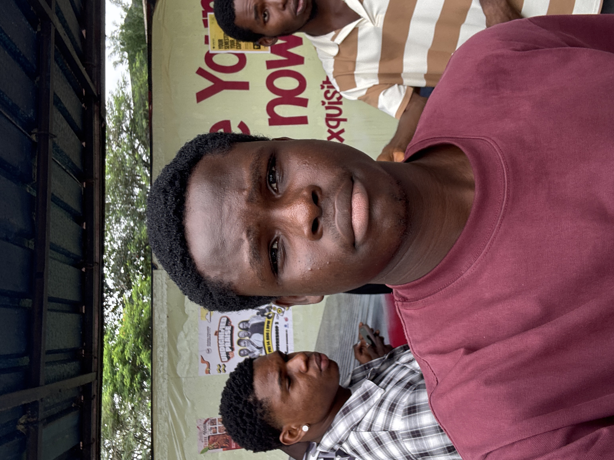 In Ihiagwa, Nigeria, a young Black male, the primary subject, is captured in a close-up selfie, facing the camera directly. He has dark, curly hair and is wearing a maroon t-shirt. To his left, another young Black male, also with dark, curly hair, is seen in profile, wearing a plaid shirt and an earring. To the right of the primary subject, partially visible, is a third young Black male wearing a striped shirt. They are positioned outdoors, in front of a yellow banner featuring red text that reads "See You now exquisite" and other smaller text. The background shows trees and a covered structure. The overall lighting suggests an overcast day.