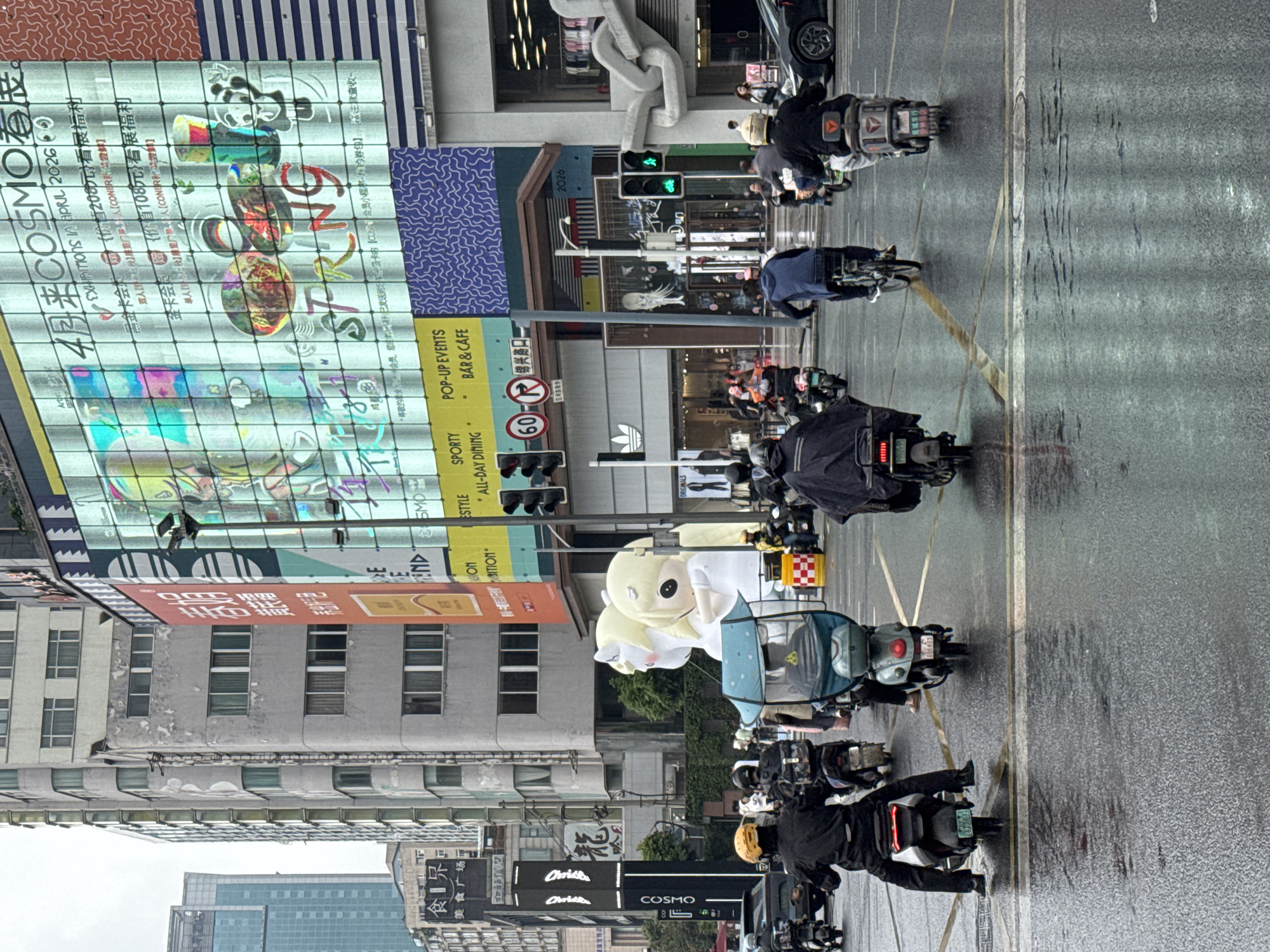 The image depicts a street scene in Jinjiang District, China, on a rainy day. A line of electric scooters with riders is moving along the road. The scooters are various colors. The riders are wearing helmets and dark clothing. In the background, there are tall buildings with advertisements on them. One building has a large "COSMO" advertisement, mentioning exhibitions in April 2026. A nearby sign has "POP-UP EVENTS BAR & CAFE" and "SPORTY ALL-DAY DINING." Street lights and traffic lights are visible. A large cartoon character or inflatable figure is secured on a scooter. There are also signs for "Christ" and "ORIGINALS."