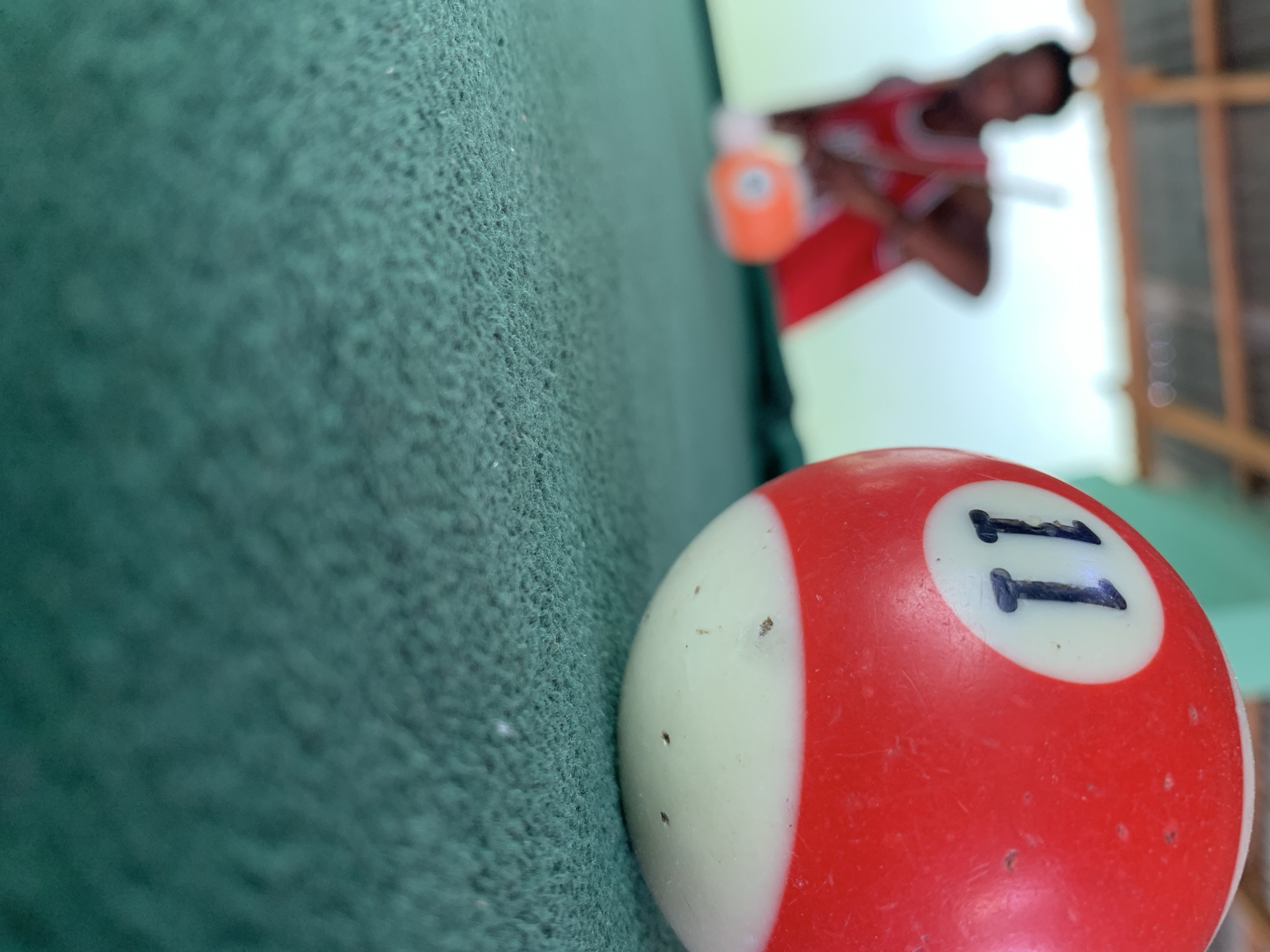 The image depicts a pool table in New Stadium, Nigeria, with a focus on a red and white striped pool ball labeled "11" in the foreground. The pool table's green felt surface is visible near the edge of the ball. In the background, a blurred figure of a person in a red shirt is visible, appearing to be playing pool or holding a pool cue. The background also shows a building or structure with wooden beams or supports.