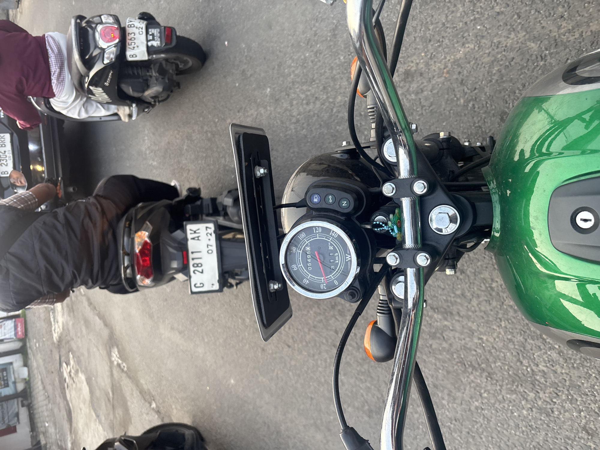 The image shows a POV perspective from a motorcycle in traffic on a paved road in West Jakarta, Indonesia. The motorcycle has a green fuel tank and a chrome handlebar with a speedometer. In front, two other motorcycles are visible, one with a rider wearing a black vest and another with a rider in a maroon jacket. A black car is in between them. The motorcycles have license plates with Indonesian alphanumeric characters. The road surface appears worn.