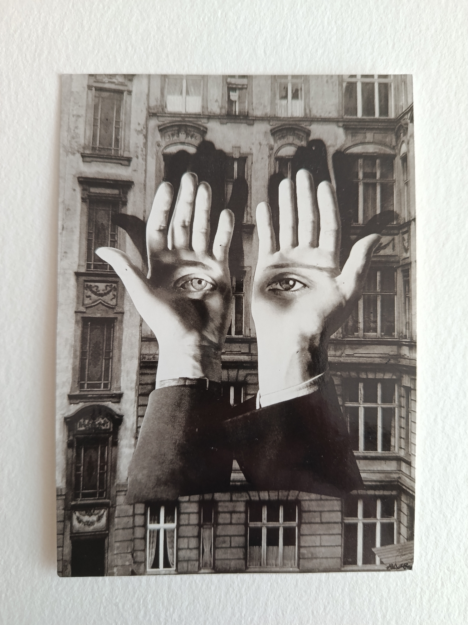 A black and white print of a surreal image is displayed against a white wall. The image features a building facade as a background. In the foreground, two hands are positioned with palms facing forward. An eye is superimposed onto the palm of each hand. The hands are attached to suit jacket sleeves, which are cut off abruptly at the bottom of the image. The print appears to be signed in the lower right corner. The building is in Moscow, Russia.