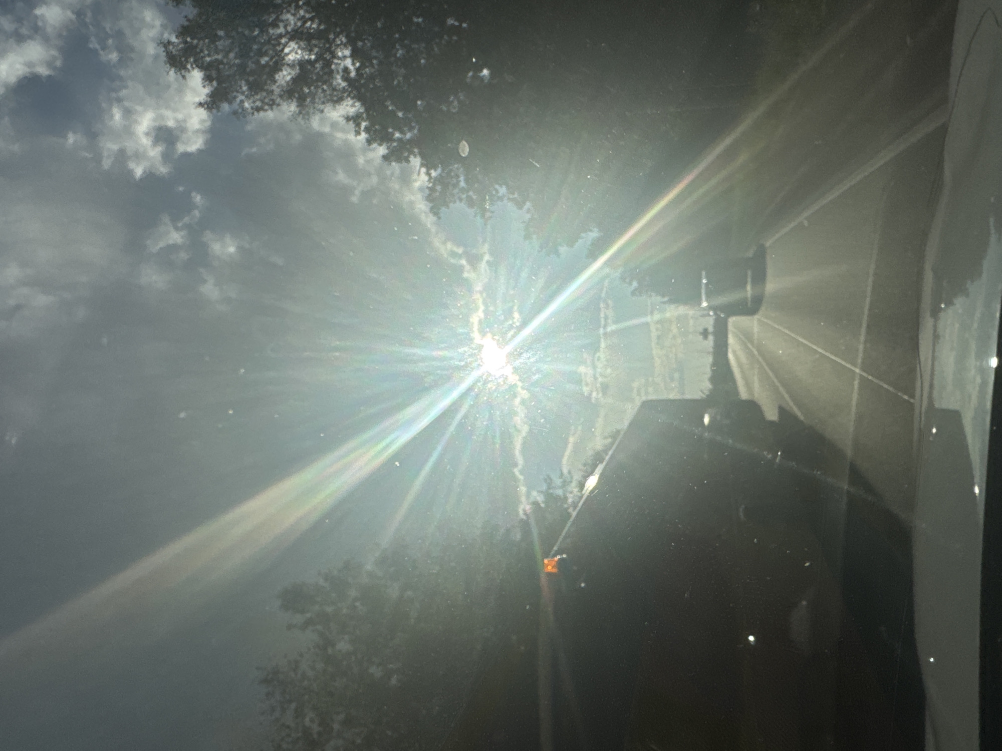 The image depicts a view from inside a vehicle traveling on a road, possibly in Dmitrievka, Russia, given the context provided. The sun is prominently featured in the sky, creating a lens flare effect with visible rays and a rainbow-like spectrum. The sky is partially cloudy. The road ahead is visible, with white lane markings. A dark-colored vehicle, possibly a truck, is in the left lane, and it has an orange flashing light on its roof. Further down the road, another vehicle is visible in the distance. Trees line both sides of the road, obscuring parts of the view. The foreground shows the interior of the vehicle from which the photo is taken, including the dashboard. The overall scene is characterized by bright sunlight and atmospheric conditions.