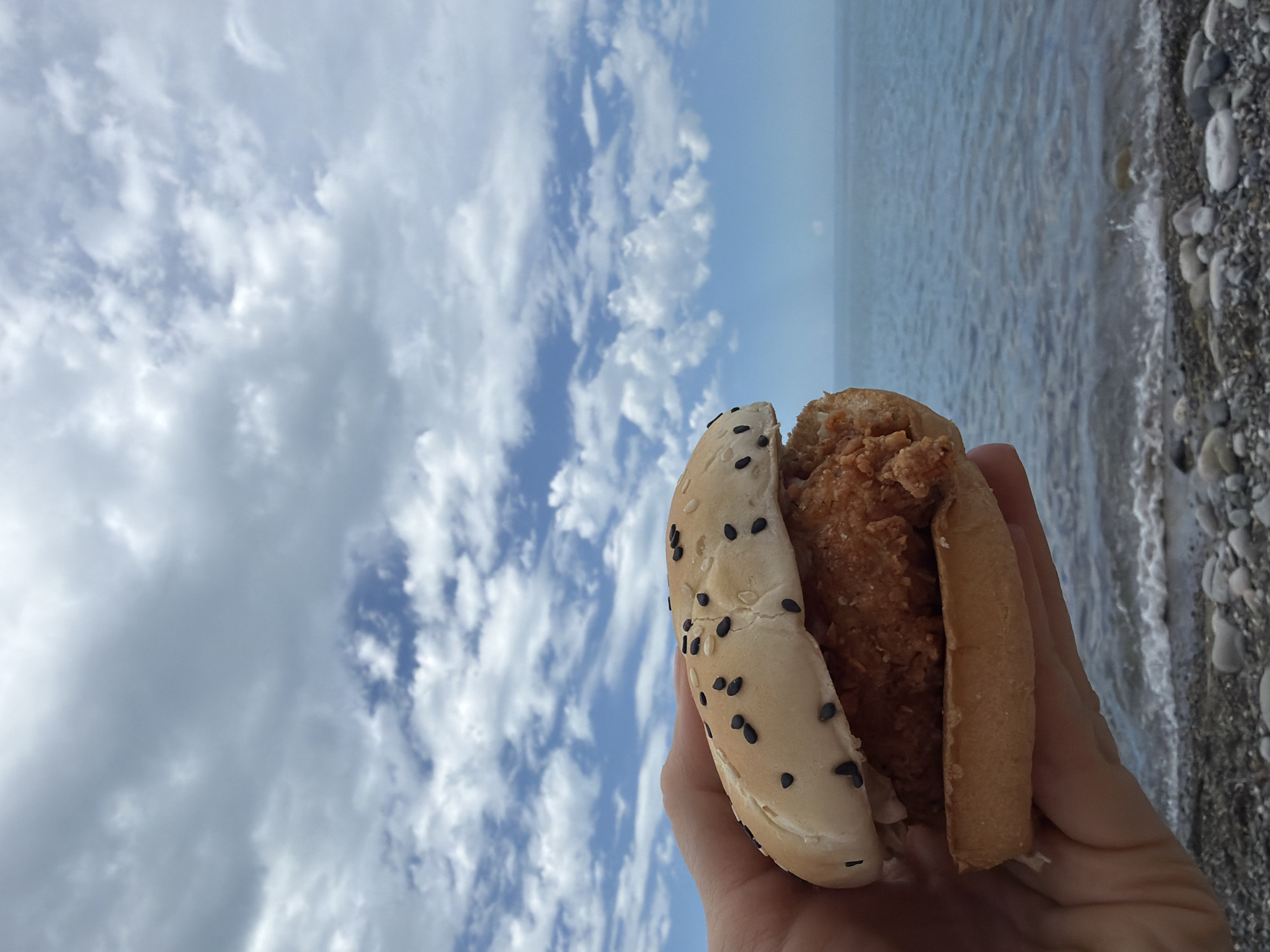 A hand holds a close-up of a fried chicken sandwich against a backdrop of the Black Sea and a cloudy sky. The sandwich has a golden-brown bun sprinkled with black sesame seeds, a thick piece of crispy fried chicken, and some white sauce peeking out. The sea is a deep blue, with small waves breaking on a pebbly beach in the foreground. The sky is filled with scattered white clouds against a pale blue expanse. The image appears to be taken on a bright, possibly overcast, day in Sochi, Russia.