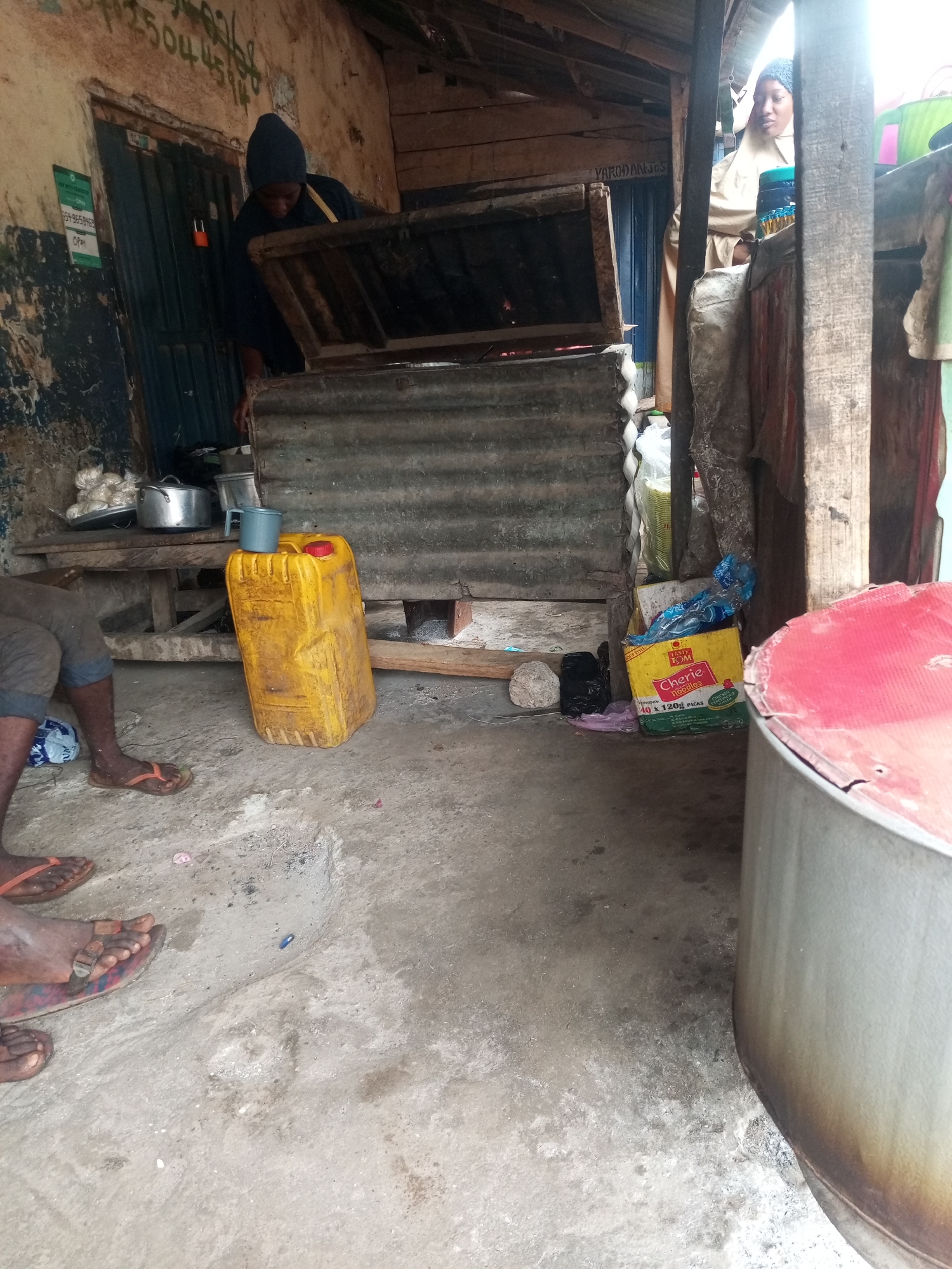 A cluttered, indoor scene in Maiburiji, Nigeria shows a food preparation area. In the background, a person wearing a dark beanie is leaning over a corrugated metal stove. Another woman, wearing a head covering and light-colored clothing, stands to the right of the stove. To the left of the stove, there is a wooden bench with metal pots and food items. In front of the stove and to the left is a large yellow plastic container with a blue mug on top. The left side of the image shows part of a person sitting on a bench wearing dark shorts and flip-flops. In the lower right corner is a large metal pot with a red lid. Behind it, a box of Cherie noodles sits next to wooden beams. The floor is concrete. The walls and beams are in disrepair. A sign on the left wall shows the phone number 125044504 and the word OPAY. There is also a name written in white paint above a door.