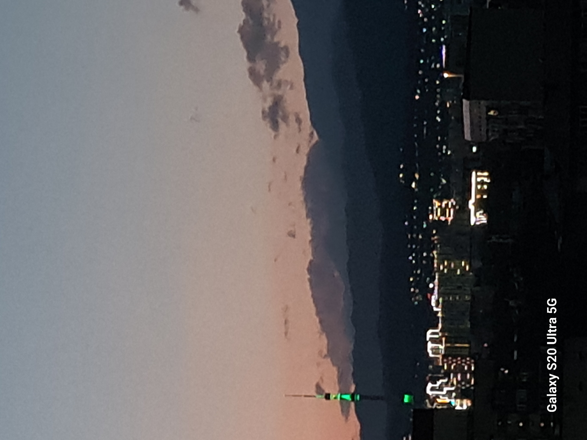 The image shows a cityscape at dusk with a prominent television tower illuminated with green lights. In the background, mountains are silhouetted against a twilight sky. The foreground features buildings with lights indicating an urban environment. The text "Galaxy S20 Ultra 5G" is present in the bottom left corner, suggesting the image was taken with this device. This scene is consistent with the view of Almaty, Kazakhstan, specifically the Kok-Tobe Tower.