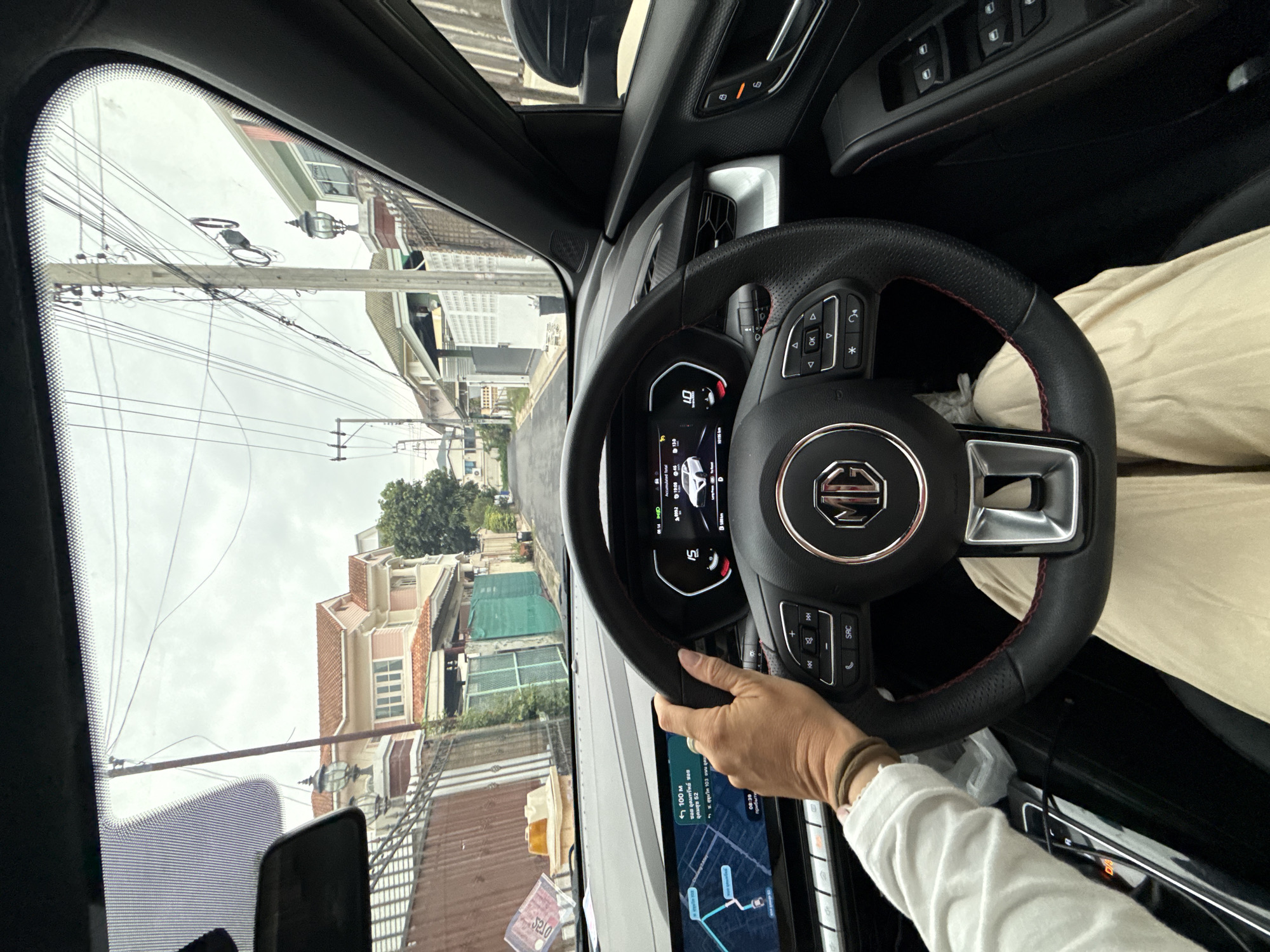 The image captures a first-person perspective from inside a car, as seen through the windshield. The driver's left hand, adorned with bracelets, is on the steering wheel, which features the MG logo. The dashboard displays a digital navigation map on the left screen and a digital instrument cluster on the right, showing a speed of 15. The car is driving down a residential street lined with houses and fences, with numerous power lines crisscrossing overhead. The sky is overcast. The houses exhibit typical suburban architecture with tiled roofs and varying color schemes. The overall environment suggests a quiet, possibly older, residential neighborhood within a city. Based on the presence of the MG car and the style of housing, this scene could plausibly be located in a suburban area of Bangkok, Thailand.