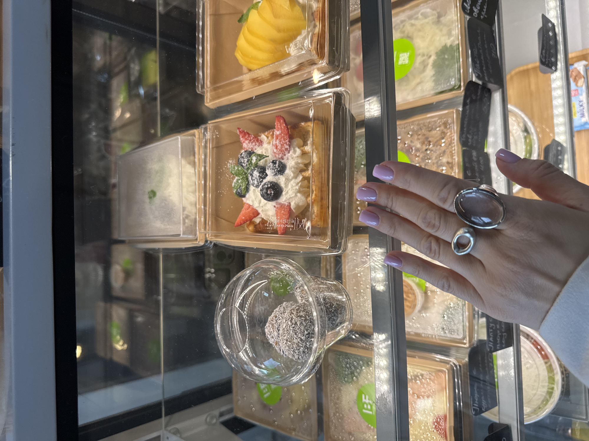 A refrigerated display case showcases a variety of desserts in individual containers. The desserts visible include a mango tart, a berry tart with whipped cream, and coconut-covered chocolate balls in a clear cup. A hand wearing two rings is visible to the right of the display. The desserts appear to be in a shop that may be located in the city of Sochi, Russia, based on the Cyrillic alphabet on a tag in the background.