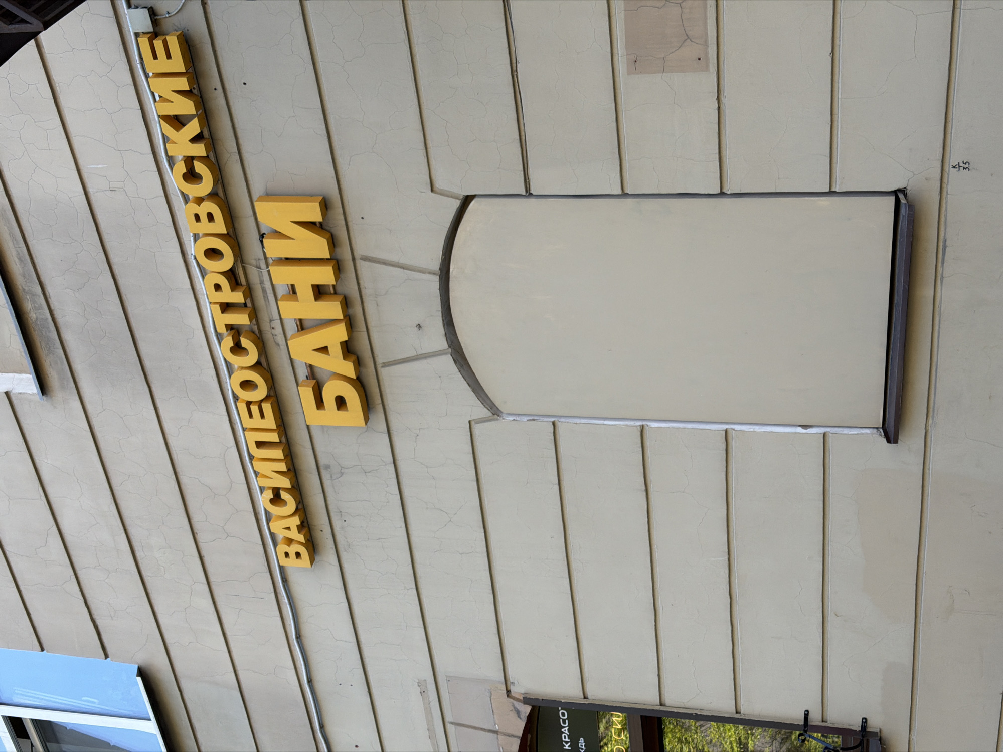 The image displays an angled, close-up view of the exterior wall of a building, likely commercial, in Saint Petersburg, Russia. The wall is clad in light-colored vertical panels, possibly stucco or siding, which are extensively covered with fine cracks, indicating age or wear.
A prominent feature is a large, raised yellow sign with Russian text, vertically oriented along the left side of the frame. The text reads "ВАСИЛЕОСТРОВСКИЕ БАНИ" (Vasileostrovskie Bani), which translates to "Vasileostrovsky Baths," referencing Vasilyevsky Island in Saint Petersburg. Electrical wires are visible running along the wall, connected to the sign.
To the right of the sign, a large, rectangular opening or alcove with an arched top has been covered or filled in with a smooth, light-colored panel. In the bottom-right corner, a part of another window or sign is visible, framed in a dark material, with partially legible Russian text that appears to show "КРАСО" (KRASO) and "ОСКИ" (OSKI). A small section of a blue-framed window is also visible in the bottom-left corner. Faint markings "x / 35" are visible on the upper-right section of the wall.
The scene is brightly lit, suggesting daytime. There are no people or explicit activities depicted, with the focus solely on the architectural details and signage.