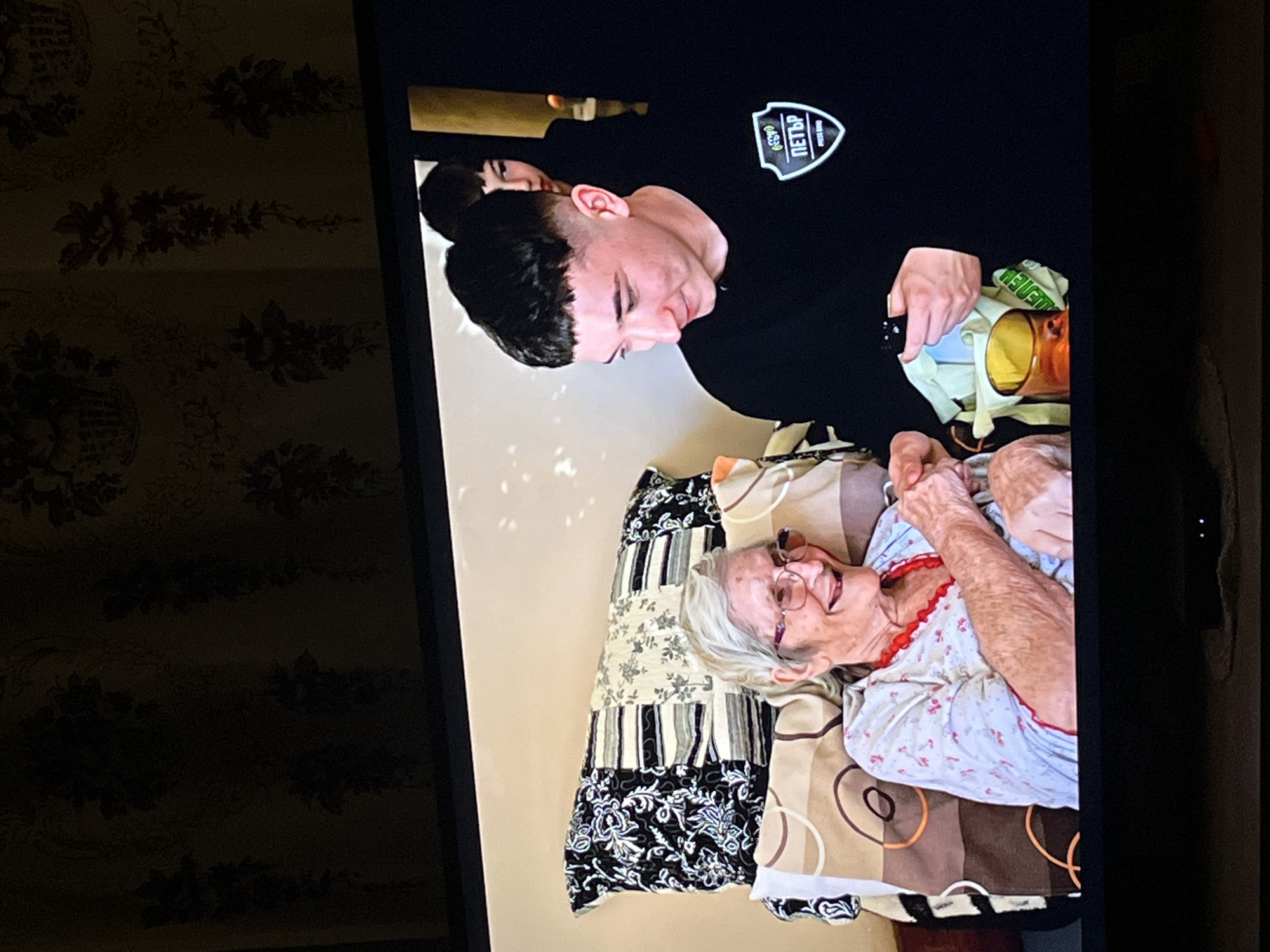 A young man in a black shirt with a patch that says "ПЕТЪР" is bending over an elderly woman lying in bed. The woman is wearing glasses and a patterned shirt with red trim. The bed has a pillow with a black and white patterned cover, and a blanket with brown and tan circles. The scene appears to be filmed on a TV screen, with a floral patterned wall and the frame of the TV visible. The location is Lozarevo, Bulgaria, based on the patch worn by the young man.