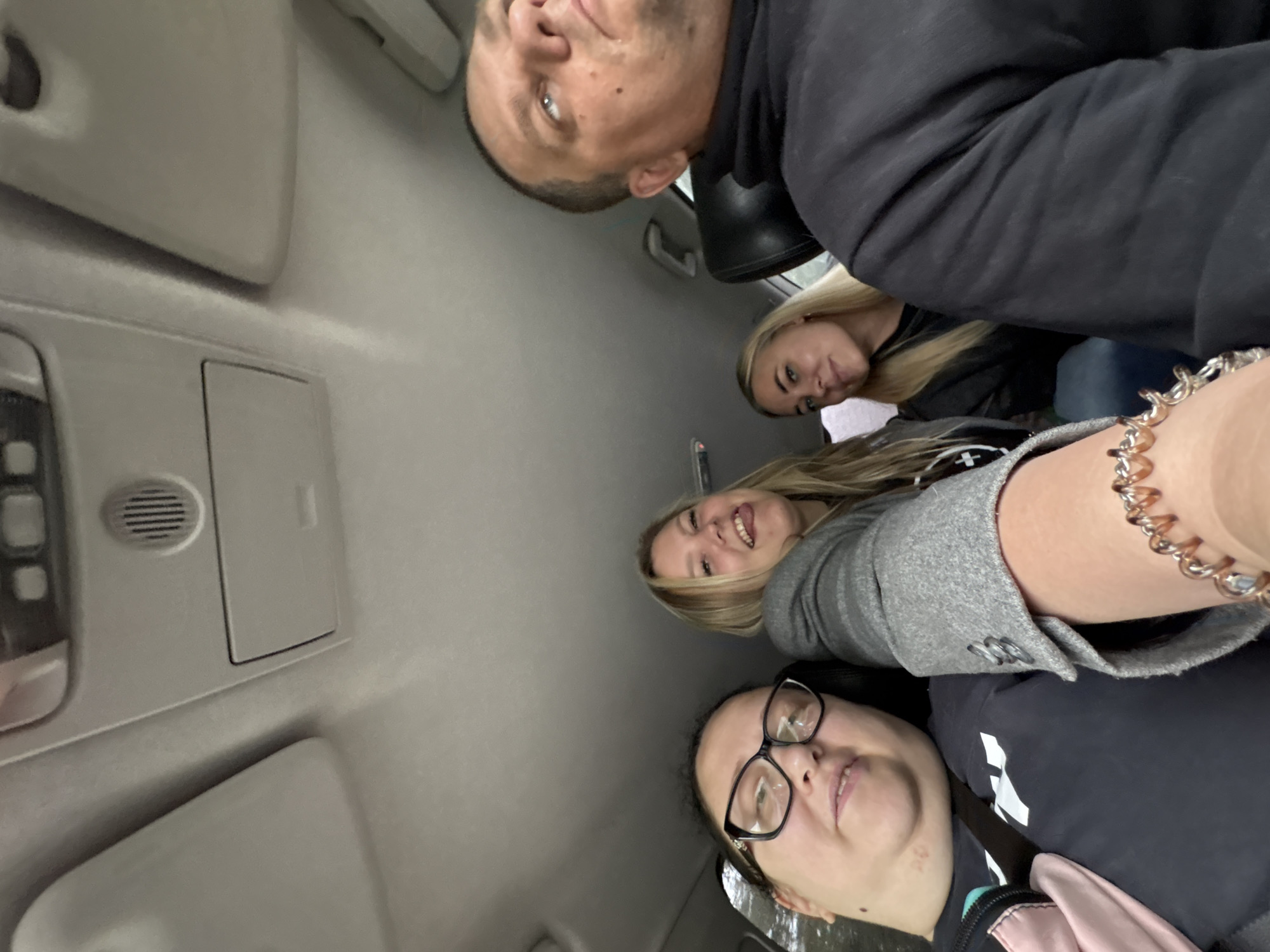 The image shows a group of four people inside a car. The camera is positioned looking up towards the occupants. One man is at the top, looking down, wearing a black jacket. Below him is a blonde woman. Another blonde woman and a woman with glasses are at the bottom of the frame. The interior of the car is visible, including the roof and some of the controls. The location is Банное, Russia.