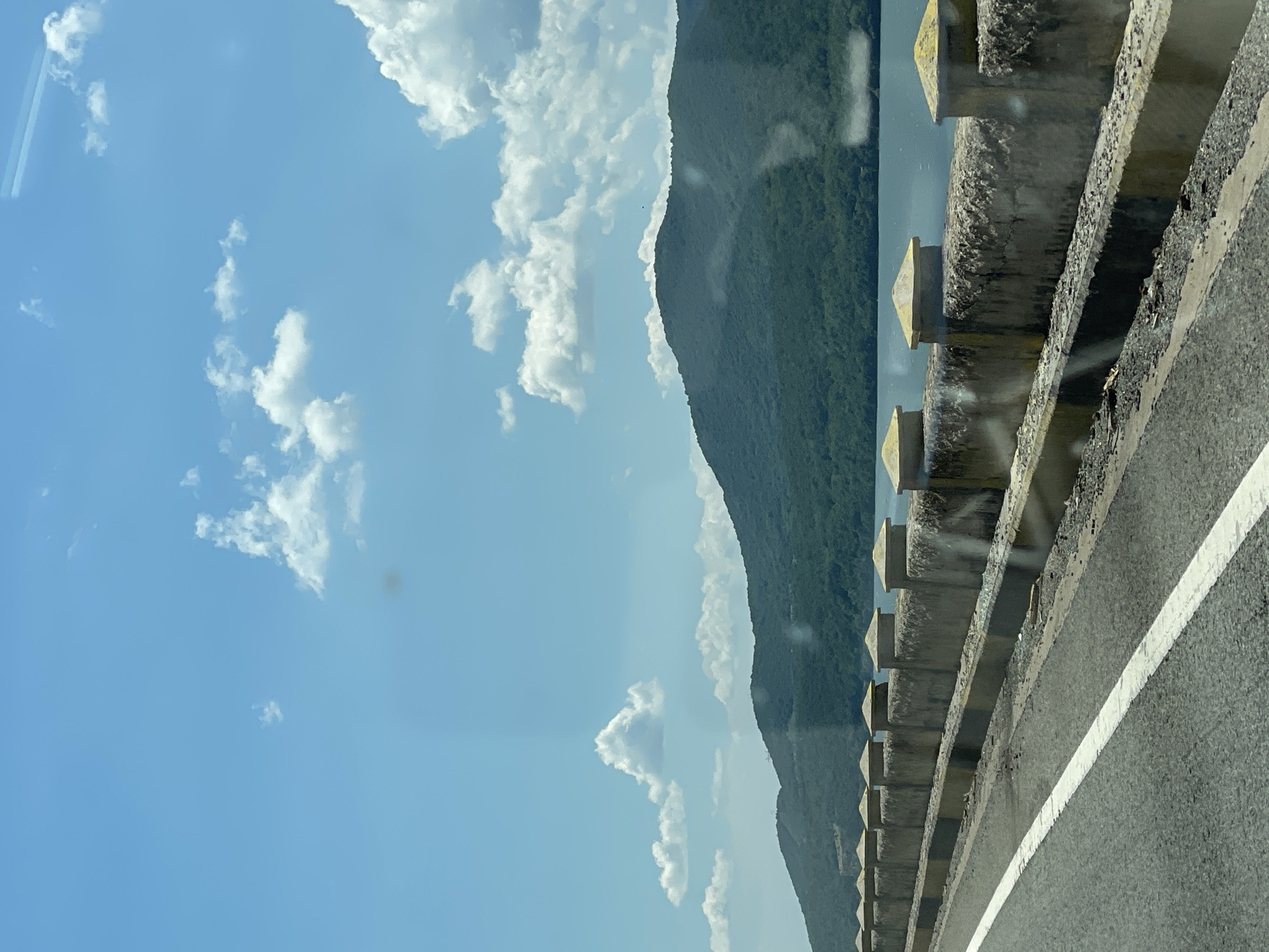 This image presents a daytime scene from a road situated alongside a large body of water, with a backdrop of forested mountains under a clear blue sky. The location is consistent with the Danube River landscape near Drobeta-Turnu Severin, Romania.
In the foreground, a grey asphalt road occupies the lower-left, featuring a worn, solid white line marking the edge of the travel lane. Parallel to the road, a concrete barrier separates it from the water. This barrier consists of a lower curb, a textured concrete wall, and a series of evenly spaced concrete posts topped with light yellow, somewhat conical caps. Debris, including small stones and litter, is visible along the base of the wall. Reflections and smudges, characteristic of a window, are apparent across the entire image, notably in the sky and water areas.
The midground features a calm, blue-grey body of water, presumably the Danube River, extending to the base of a series of lush, green, densely forested mountains.
The background is dominated by the undulating profiles of these mountains, covered in extensive tree canopy. Above the mountains, a bright blue sky contains numerous scattered white cumulus clouds of varying sizes. A small, dark, indistinct object, possibly avian, is visible high in the sky above the mountain range. No identifiable individuals or direct interactions are discernible within the frame.