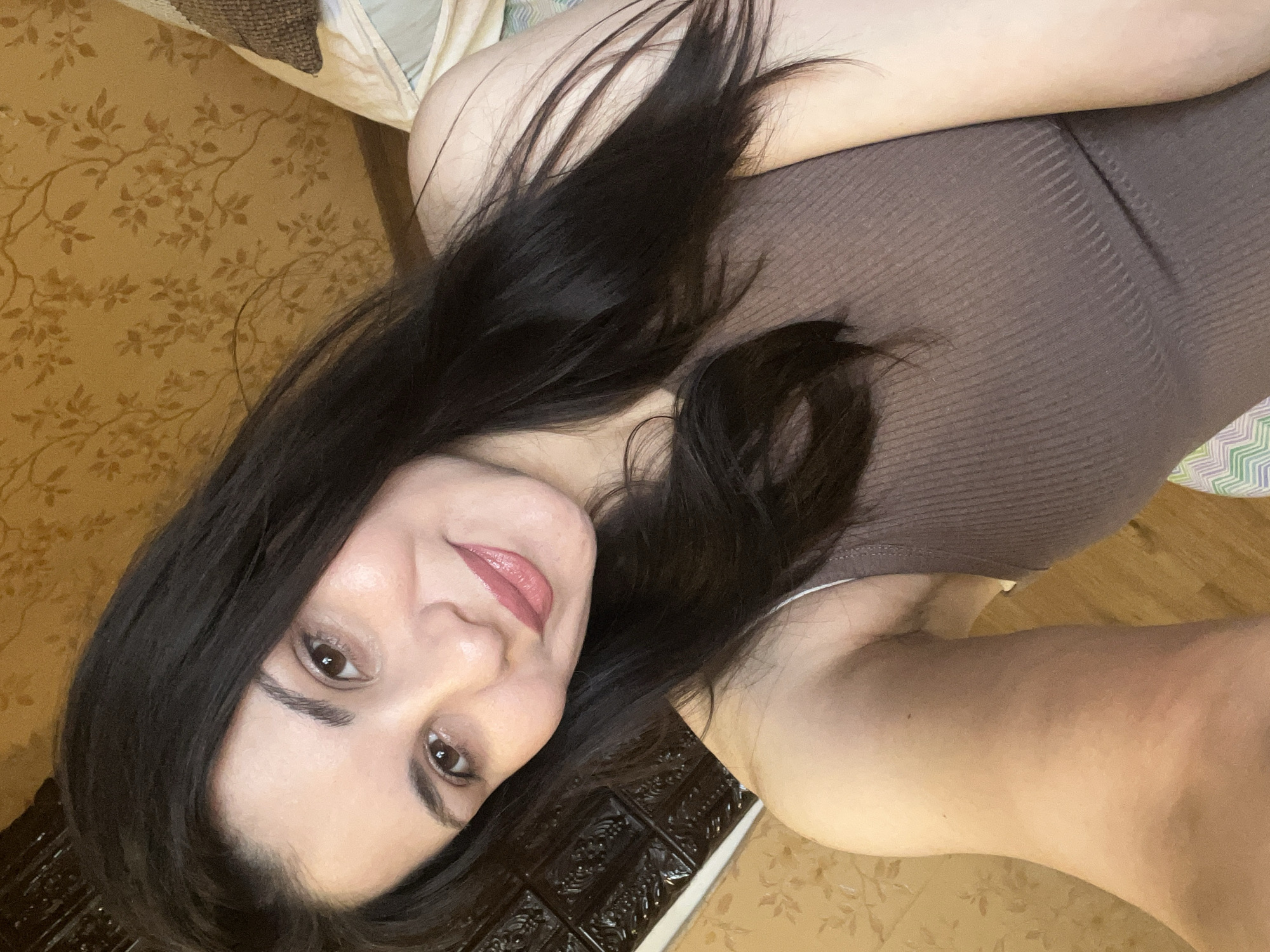 A young woman with long, dark hair and brown eyes takes a selfie indoors. She is wearing a brown ribbed tank top and has light-colored lipstick on. Her skin appears smooth and radiant. She is angled slightly away from the camera, with her left shoulder visible. The background shows a wall with floral wallpaper and a dark, textured panel. A pillow and what appears to be bedding are also visible. The lighting is warm and soft, suggesting an indoor setting.