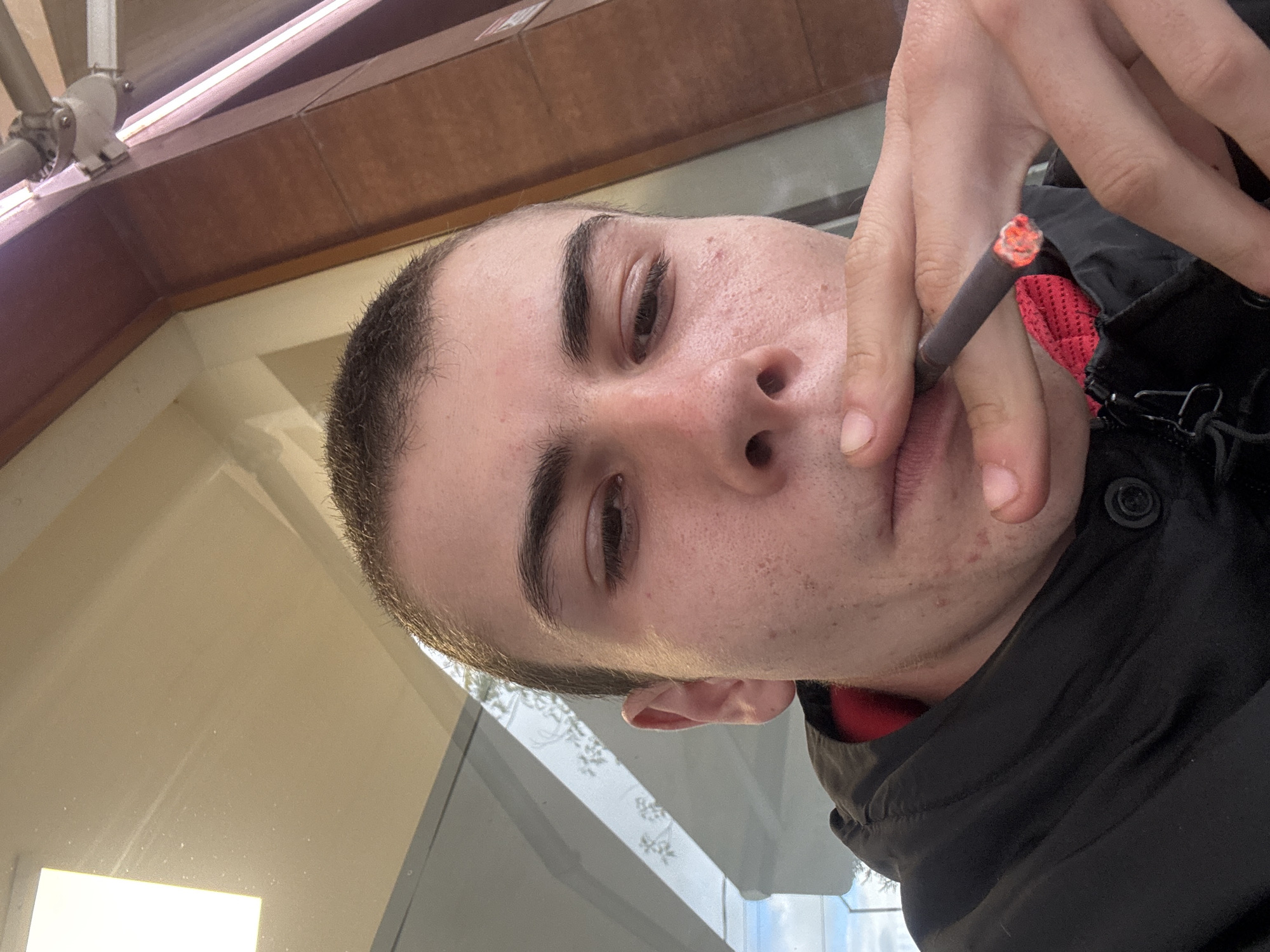 A person is holding a lit cigarette to their lips. The background shows a building exterior and what appears to be a window, suggesting an outdoor or semi-outdoor location. The setting may be Plovdiv, Bulgaria. The individual is wearing a dark jacket with a button near the collar and has short hair. The cigarette is partially obscured by the person's fingers.