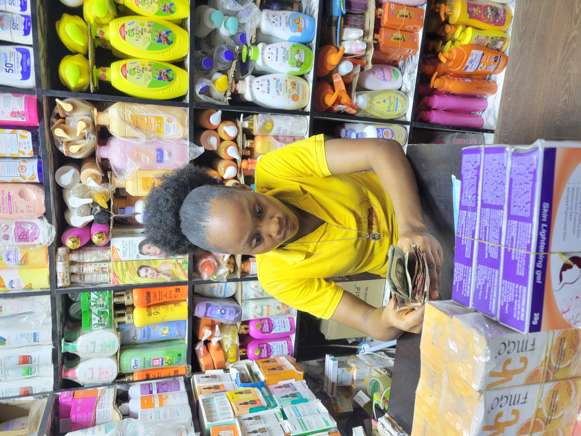 Here is a detailed description of the image:
**Scene:**
The image shows a small retail space or shop, possibly in Onitsha, Nigeria, based on the products and currency. The space is filled with shelves packed with various personal care and cosmetic products.
**People:**
A woman with dark hair pulled back into a ponytail, wearing a bright yellow shirt, is the main subject. She is positioned behind a counter and is shown counting a wad of Nigerian currency.
**Subjects:**
The focus is on the woman counting money amidst a store filled with products.
**Objects:**
* **Products:** Shelves are laden with a wide variety of products: lotions, body washes, creams, and gels. Brands like "Clere," "Dove," "Johnson's," "Feminelle," "FMGC" and others are visible.
* **Currency:** The woman is counting a stack of Nigerian Naira notes.
* **Shelving:** The products are arranged neatly on dark-colored wooden shelves.
* **Packaging:** Many items are in colorful plastic bottles or cardboard boxes. One box is labelled as "Skin Lightening Gel."
**Actions:**
The woman is actively counting money.
**Interactions:**
The woman is focused on counting the money, seemingly handling a transaction.
**Environment:**
The setting is a small, cluttered retail space. The environment suggests a busy commercial area.