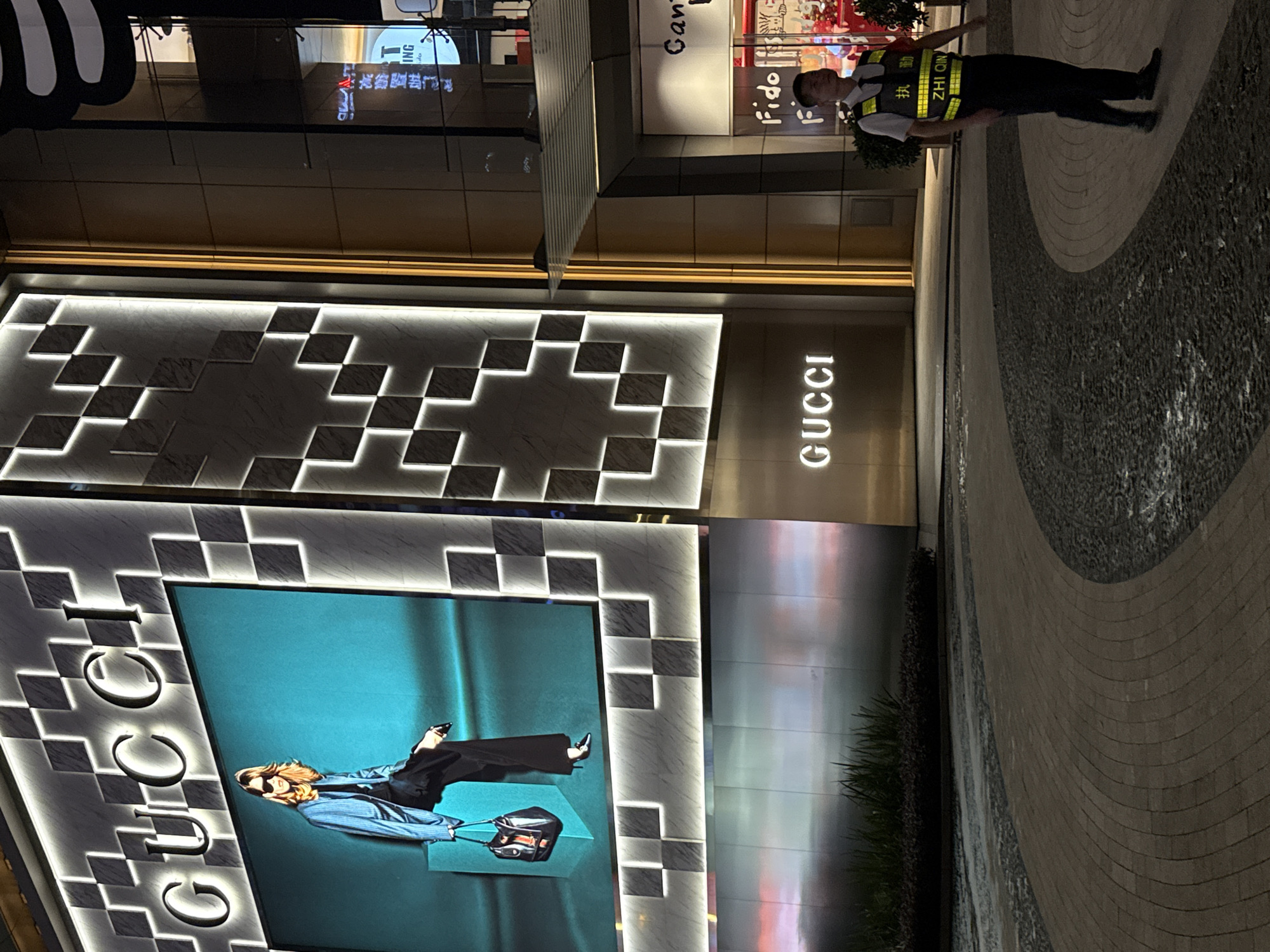 The image depicts an outdoor scene at night in Tianhe District, China, likely a commercial area. The primary subject is a security guard wearing a white shirt, black pants, and a high-visibility yellow vest with reflective stripes. The vest has Chinese characters on it, reading "执勤" (zhí qín) and "ZHI QIN". He is standing on a patterned stone walkway.
Behind the guard, a large building facade is visible, featuring the illuminated "GUCCI" logo in prominent white lettering. The building's design incorporates geometric patterns with integrated lighting. A large digital screen displays an advertisement featuring a model in a blue jacket and dark pants, posing with a handbag. The model is wearing sunglasses and has blonde hair.
The overall setting appears to be a modern urban environment, possibly a shopping mall or a high-end retail street. The lighting suggests it is nighttime, and the well-maintained pavement and illuminated signage indicate a commercial or entertainment district. The scene is well-lit, creating a vibrant atmosphere despite the late hour. The security guard appears alert and observant, performing his duties. The advertisement on the screen is a clear indicator of luxury retail.