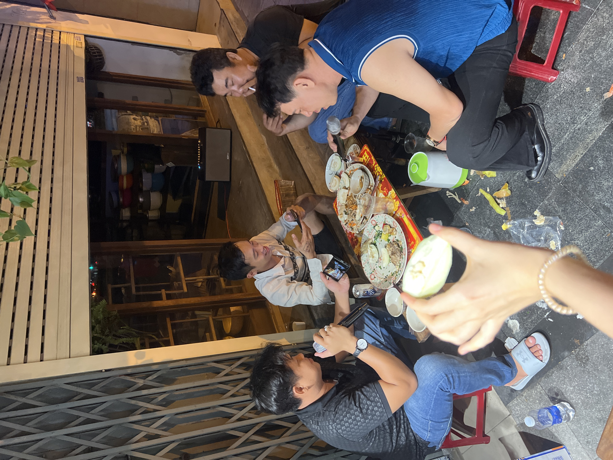 The image depicts a nighttime street scene in the city of Thủ Đức, Vietnam. Five men are gathered around a low table laden with food scraps, empty plates, and beverage containers. One man is smoking a cigarette, while another holds a microphone. A third man appears to be filming or taking a picture with his phone. One person holds a halved cucumber, possibly preparing to eat it. The men are dressed in casual clothing. A metal security gate is visible in the background. A water bottle lies on the pavement.