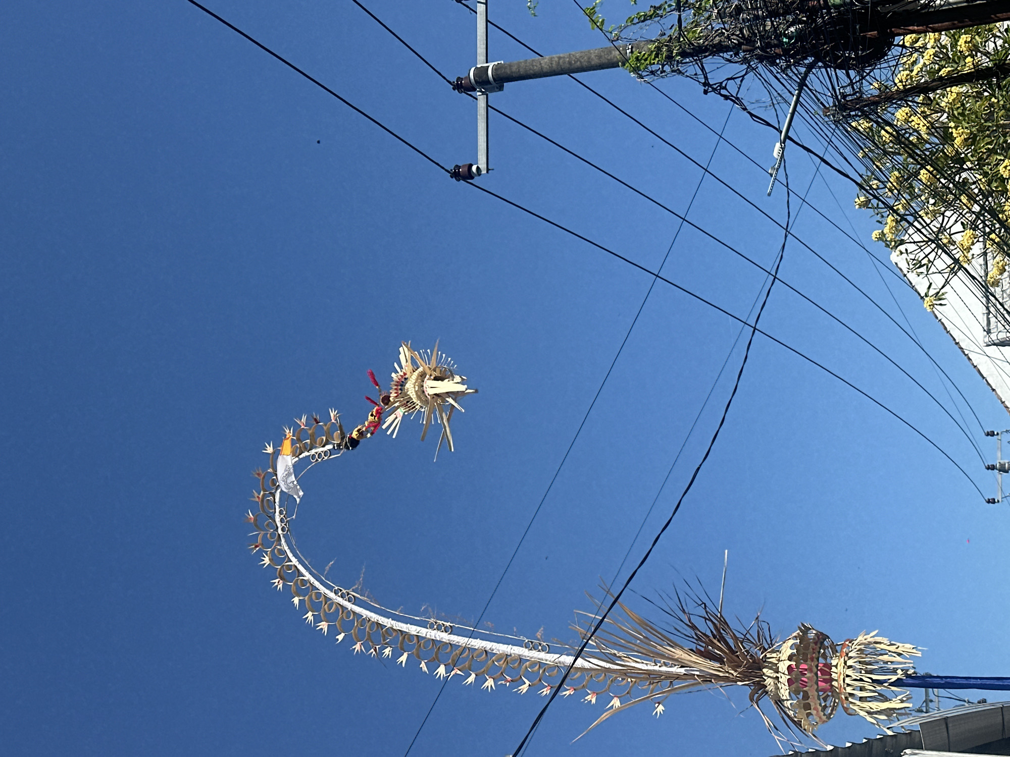 This is a vertical, medium shot taken from a low angle, looking up at a clear blue sky with a decorative Balinese penjor on the left and a utility pole on the right. The penjor is a tall, arched bamboo pole adorned with woven palm leaves, flowers, and small flags, extending from the bottom left of the frame upwards and curving to the right. The upper section of the penjor features a more elaborate decoration resembling a dragon's head made of wood and straw, with colorful fabric accents. Wires from the utility pole crisscross the sky, some connected to the pole which is partially covered in green vines. Below the pole, the top of a white building and yellow flowers are visible. The scene is likely in North Kuta, Indonesia, judging by the presence of the penjor, a common Balinese offering. There are no people or discernible actions taking place.