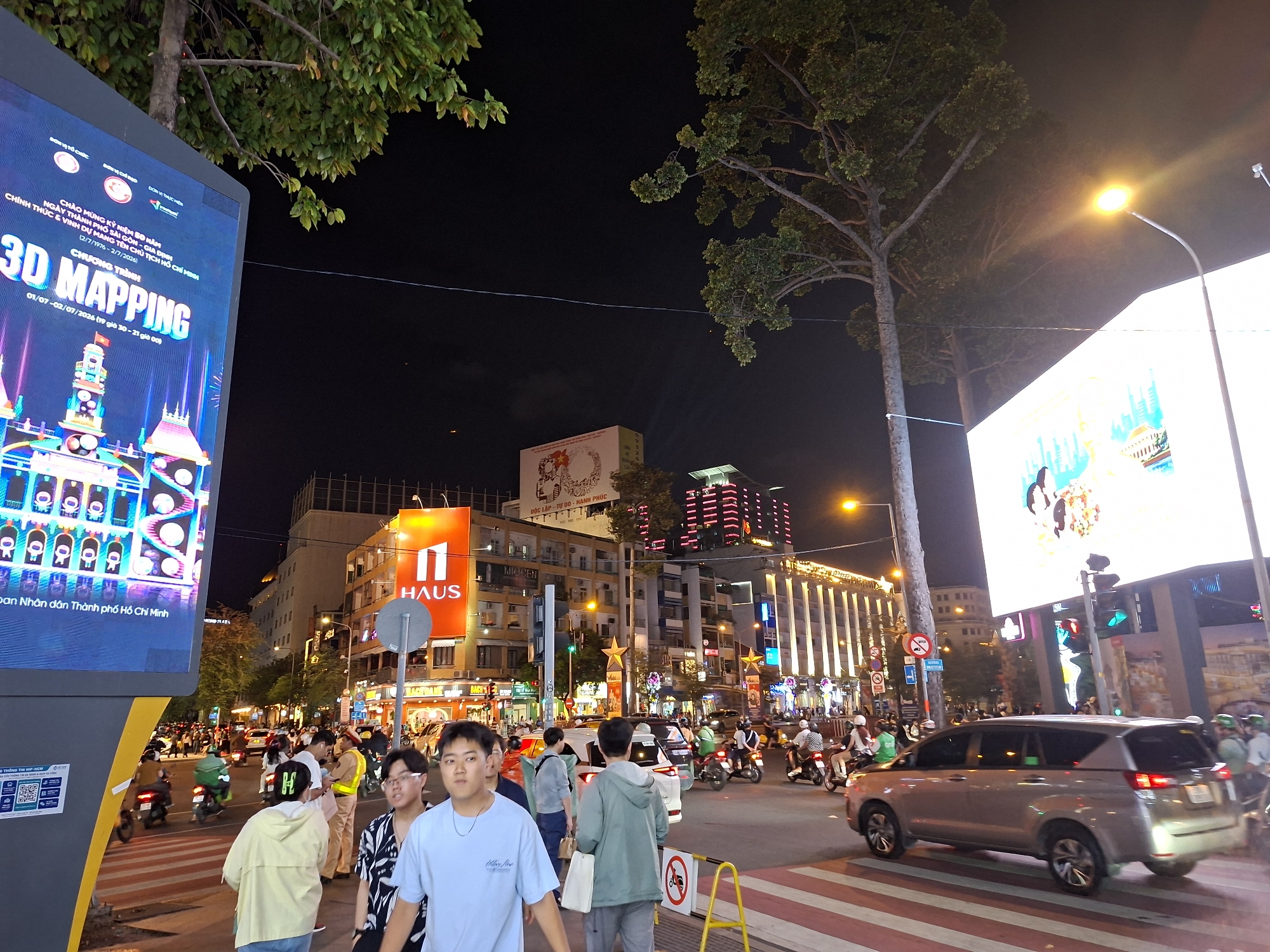 This image captures a nighttime street scene in Thủ Đức City, Vietnam, likely a commercial or entertainment district. A large digital billboard on the left displays "3D MAPPING" with colorful imagery of buildings. Across the street, a bright "HAUS" sign illuminates a building. The street is busy with traffic, including numerous motorcycles, cars, and a silver SUV in the foreground. Pedestrians are visible on the sidewalk and crossing the street at a marked crosswalk. Traffic lights are visible, with one showing a red light. The street is lined with multi-story buildings adorned with lights and advertisements. Trees with foliage frame the upper part of the image, and a street lamp provides overhead illumination. The overall atmosphere is lively and urban, indicative of a bustling city at night.