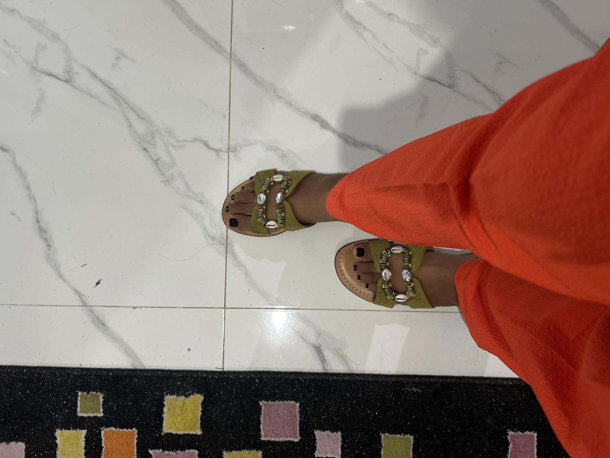 The image shows a person's feet wearing open-toed sandals with green straps adorned with shells and clear stones, against a background of white marble tile flooring. The person is also wearing loose-fitting orange trousers. To the left of the frame, a black rug with colorful square patterns is visible. The scene appears to be indoors. There is no specific information that allows for the determination of the location within Sukawati, Indonesia.