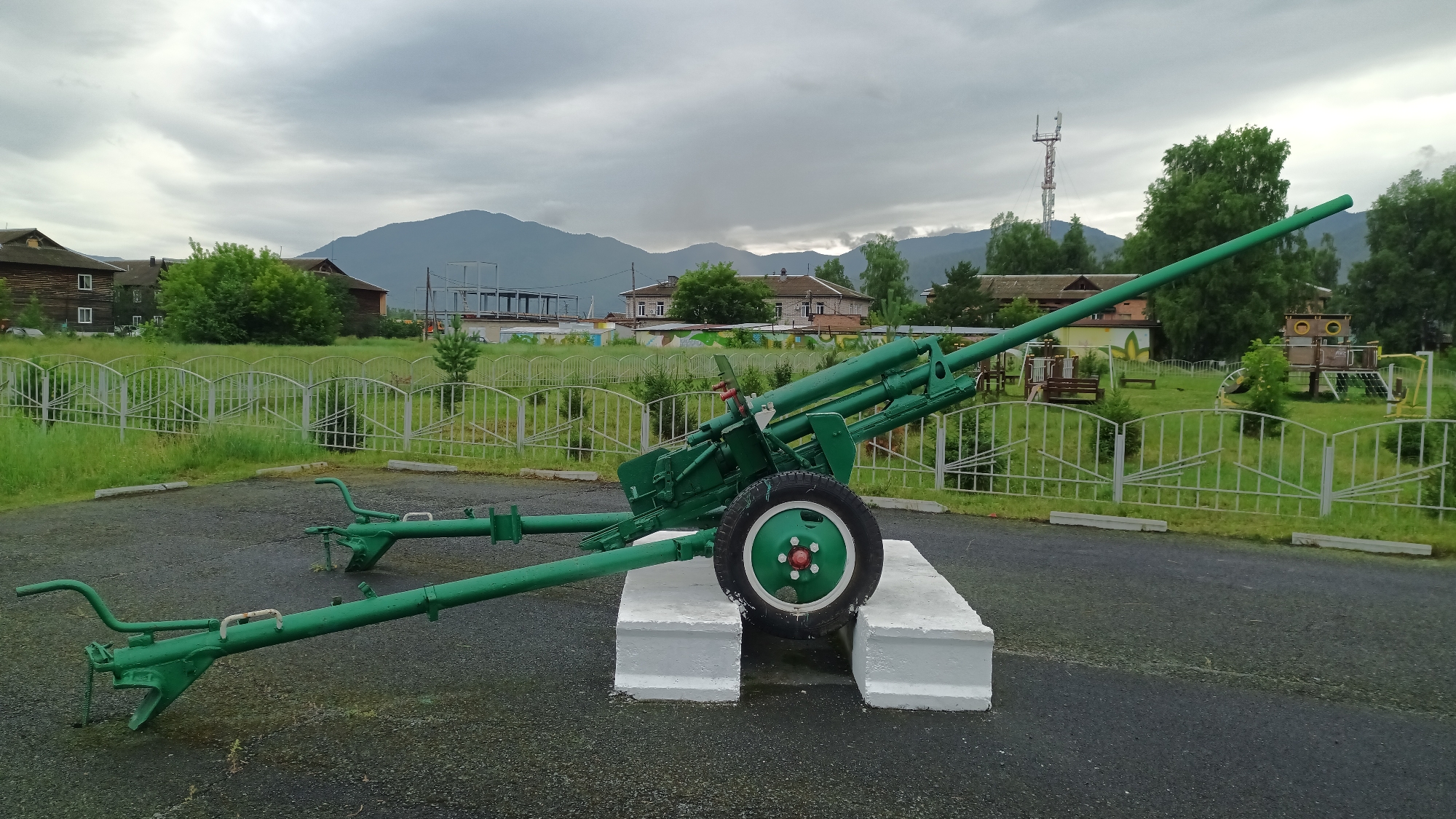 The image captures a bright green artillery piece displayed outdoors in what appears to be a park or public area. The cannon is positioned on two white concrete blocks, with its barrel extending diagonally across the frame from left to right, pointing towards the right. The artillery piece has a single wheel with red accents on its hub. Extending from the rear of the cannon are two legs, also painted green, which are spread outwards.

In the background, a white metal fence with a decorative pattern encloses a grassy area. Beyond the fence, several buildings are visible. Some appear to be made of dark wood, suggesting a rustic or traditional architectural style. Others are more modern, with tiled roofs. Lush green trees and foliage are scattered throughout the scene, and in the far distance, a range of mountains under a cloudy sky forms the backdrop. A tall communication tower with antennas is also visible on the horizon.

The sky is overcast, indicating a cloudy or potentially rainy day, and the overall lighting is diffused, suggesting it is daytime. The ground in the foreground is dark and appears to be wet, consistent with recent rain. The setting seems to be a small town or village, as suggested by the mix of residential buildings and the presence of a public space. There are no people visible in the image, and the mood is neutral, with the focus solely on the static display of the military artifact within its environment. The location is identified as Abaza, Russia.