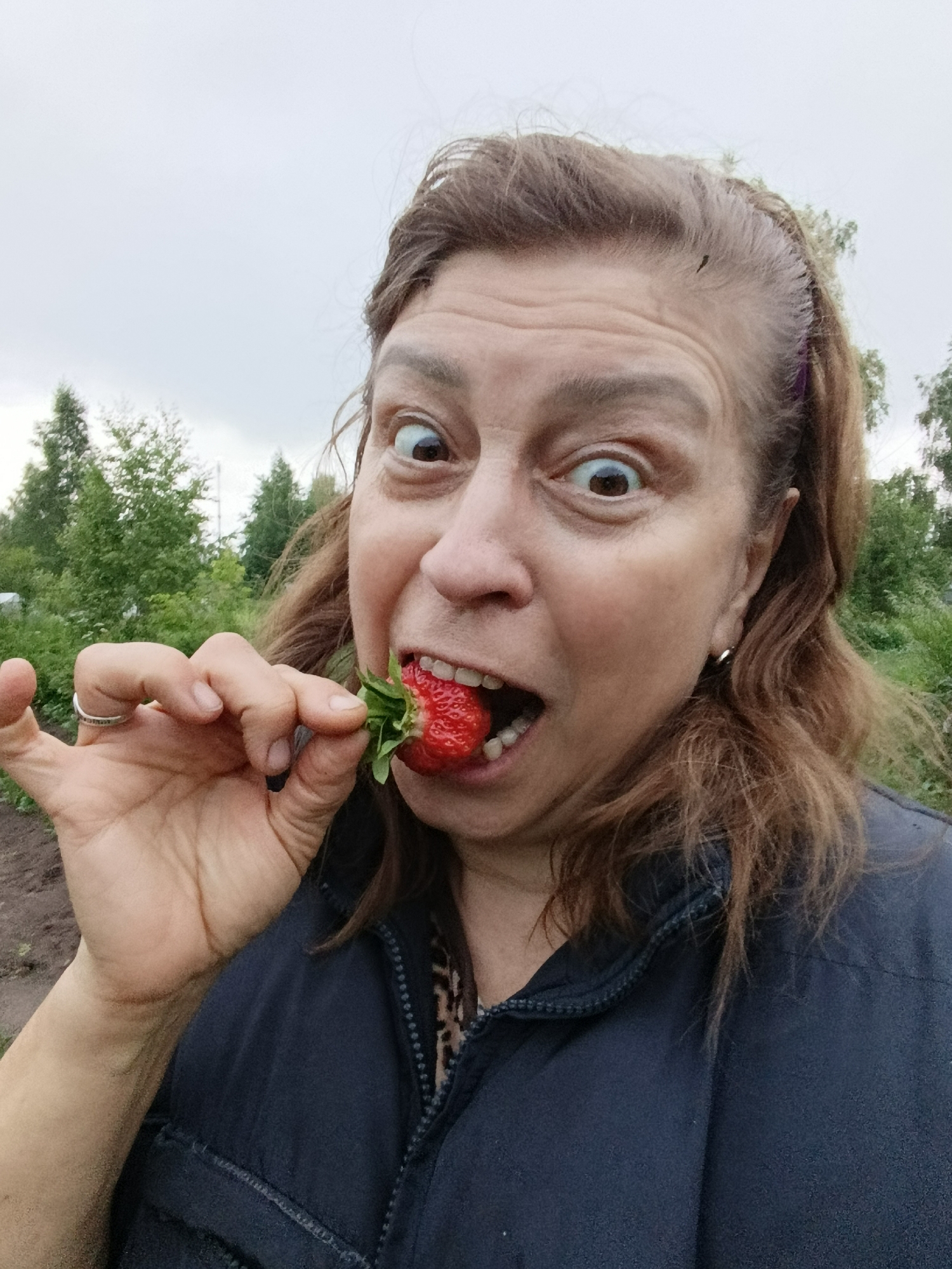 A woman is taking a selfie outdoors, holding a strawberry to her mouth. She has brown hair, wide eyes, and is wearing a dark blue jacket over a leopard-print top. She is also wearing a ring on her left hand. The background shows trees and an overcast sky. The location is likely in the city of Vereya, Russia, suggested by the presence of trees and an outdoor, possibly rural or garden setting.