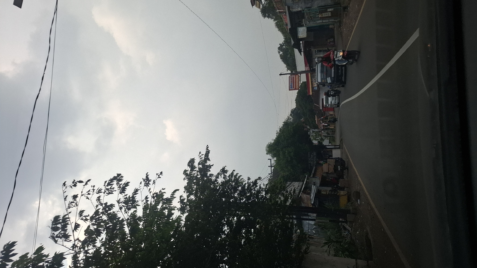 The image depicts a street scene in Cilaku, Indonesia, under an overcast sky. On the left, a large tree with dark green foliage overhangs the paved road. Along the side of the road, there are several small buildings and stalls, some with awnings and wares displayed. A dark-colored vehicle is parked near one of the buildings.
The road itself has a white dividing line. Several vehicles are present: a red motorcycle with a rider wearing a red jacket and a helmet is in the foreground on the right side of the road, moving away from the viewer. A white van is further up the road on the left, and a black pickup truck is also visible. Another motorcycle is positioned behind the white van. A sign for "Alfamart" is visible in the background, indicating a commercial area. The environment appears to be a mix of residential and commercial properties, with power lines running across the sky. The overall impression is of a typical day on a local road in Indonesia.