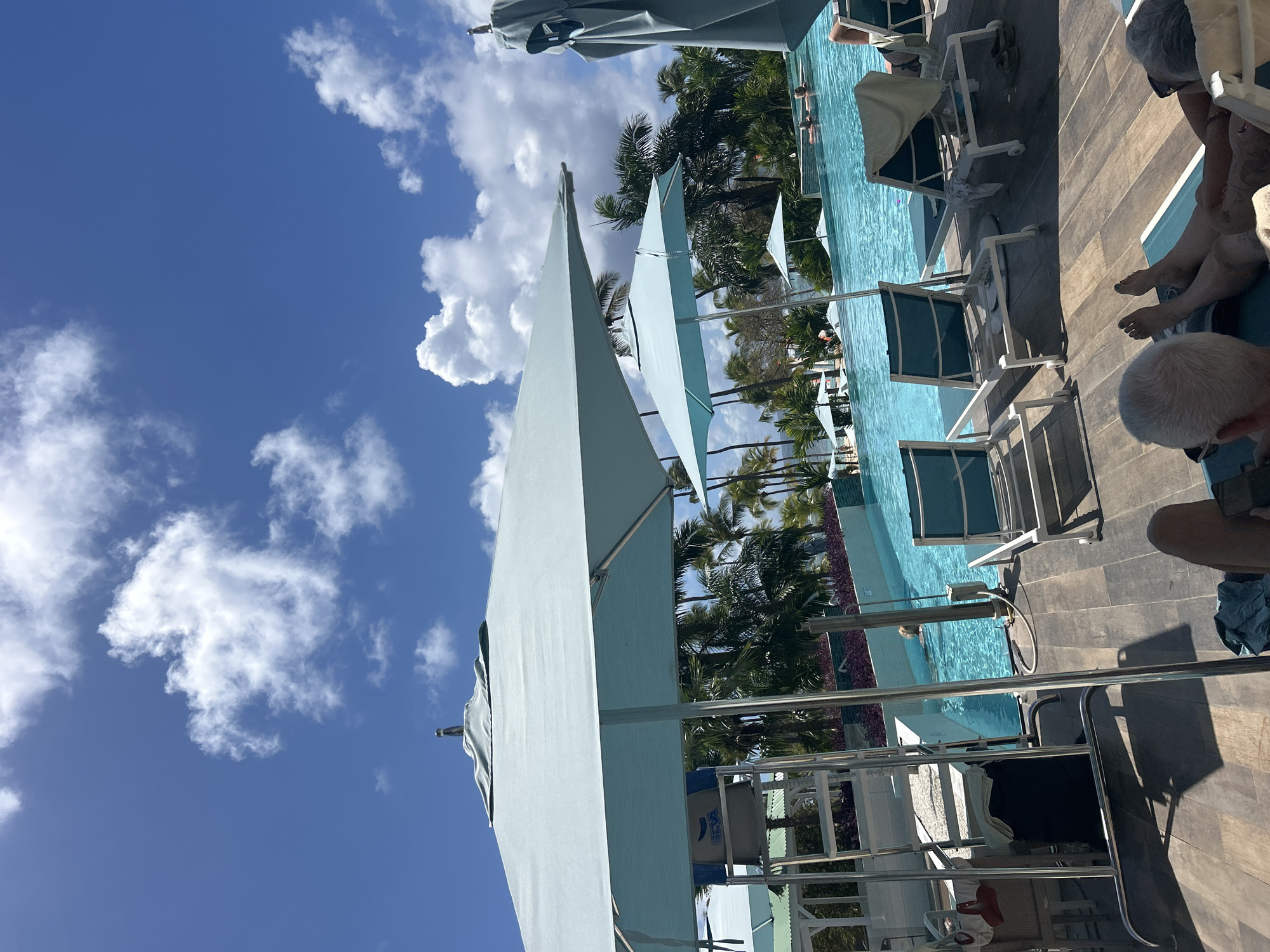 This image captures a wide-angle view of a resort swimming pool area in Sainte-Anne, France, under a clear blue sky with scattered white cumulus clouds. In the foreground, two individuals are visible, reclining on light-colored lounge chairs with teal cushions on a slatted wooden deck. The person on the left displays the back of their head with short grey hair, while the person on the right shows their bare feet and lower legs.
Multiple large, light-blue rectangular shade umbrellas are open, providing shade over the deck and pool. One umbrella dominates the left foreground, obscuring part of a tall, silver-framed lifeguard chair positioned by the pool's edge, which has a beige bag with blue lettering ("CEVA") hanging from its back. A large, turquoise-colored swimming pool occupies the midground, with several individuals partially visible in the water, appearing to be swimming or wading.
The background features dense tropical vegetation, including numerous palm trees and other lush greenery, consistent with a resort environment. Distant white-colored resort buildings with light roofs are partially visible through the trees. The overall scene depicts a sunny, leisurely outdoor setting.
