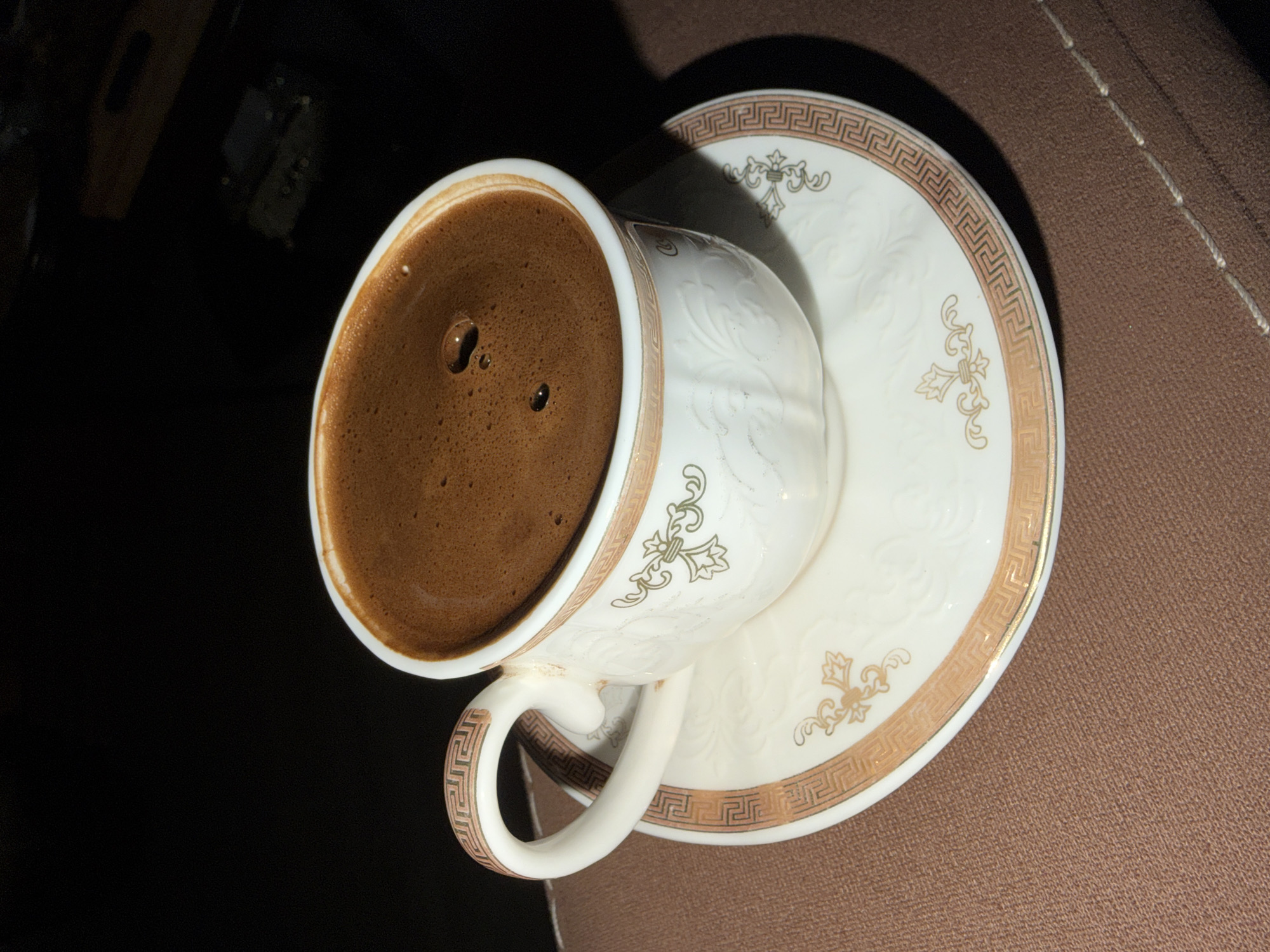 A cup of dark-brown coffee sits on a white and gold saucer. The cup is also white and gold and has a raised design. The coffee is nearly to the rim. The saucer is resting on a brown surface, possibly fabric. The background is dark, with a dim reflection visible in the upper-left corner. The location is not discernible from the provided image and therefore, cannot be linked to Krasnoyarsk, Russia.