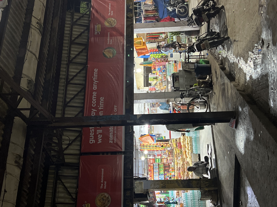 This night-time image captures a busy street scene in Palani, India, under artificial lighting. The left side of the frame is dominated by a dark corrugated metal roof supported by robust metal beams, suggesting an overhead structure like a canopy or part of a market building.
Below this structure, a series of brightly lit shops line the street. These shops display various products, including shelves stocked with bottled beverages and other general merchandise. Large stacks of bottled water are visible outside one of the establishments.
Several people are present in the scene. In the foreground on the right, a person is seated on the ground or a low stool. Further back, inside one of the shops, another person stands. Multiple motorcycles are parked on the right side of the street, and a bicycle is also visible near one of the shops. The ground appears damp in places, reflecting the light from the shops and streetlights.
Prominently displayed are several large red advertising banners. Visible text on these banners includes "The Annavasal Hotel" (appearing twice), "guest may come anytime we'll on time," and "zomat delivers on tim % OFF," which likely refers to the food delivery service Zomato. Another banner features the word "TRENDING" and "Grand Grooms Hotel." The scene portrays a lively commercial area, likely late in the evening or at night.