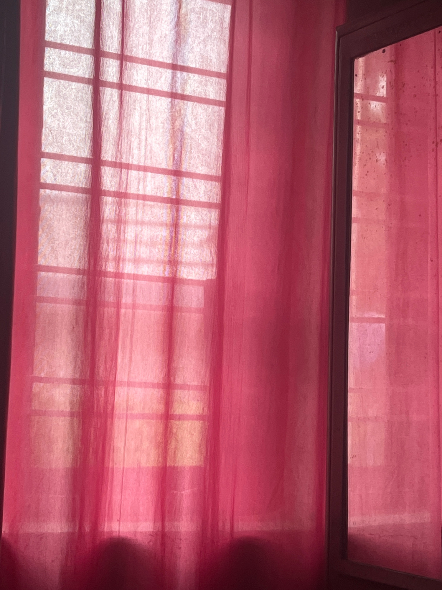 The image shows a window with pink curtains in Chokandalli, India. The curtains are sheer, allowing light to filter through and reveal the grid pattern of the window. The right side of the image shows a reflection in what appears to be a mirror or another window, also with pink curtains and a similar grid pattern. The lighting suggests it is daytime. There are no people or explicit actions depicted in the image. The environment appears to be indoors.