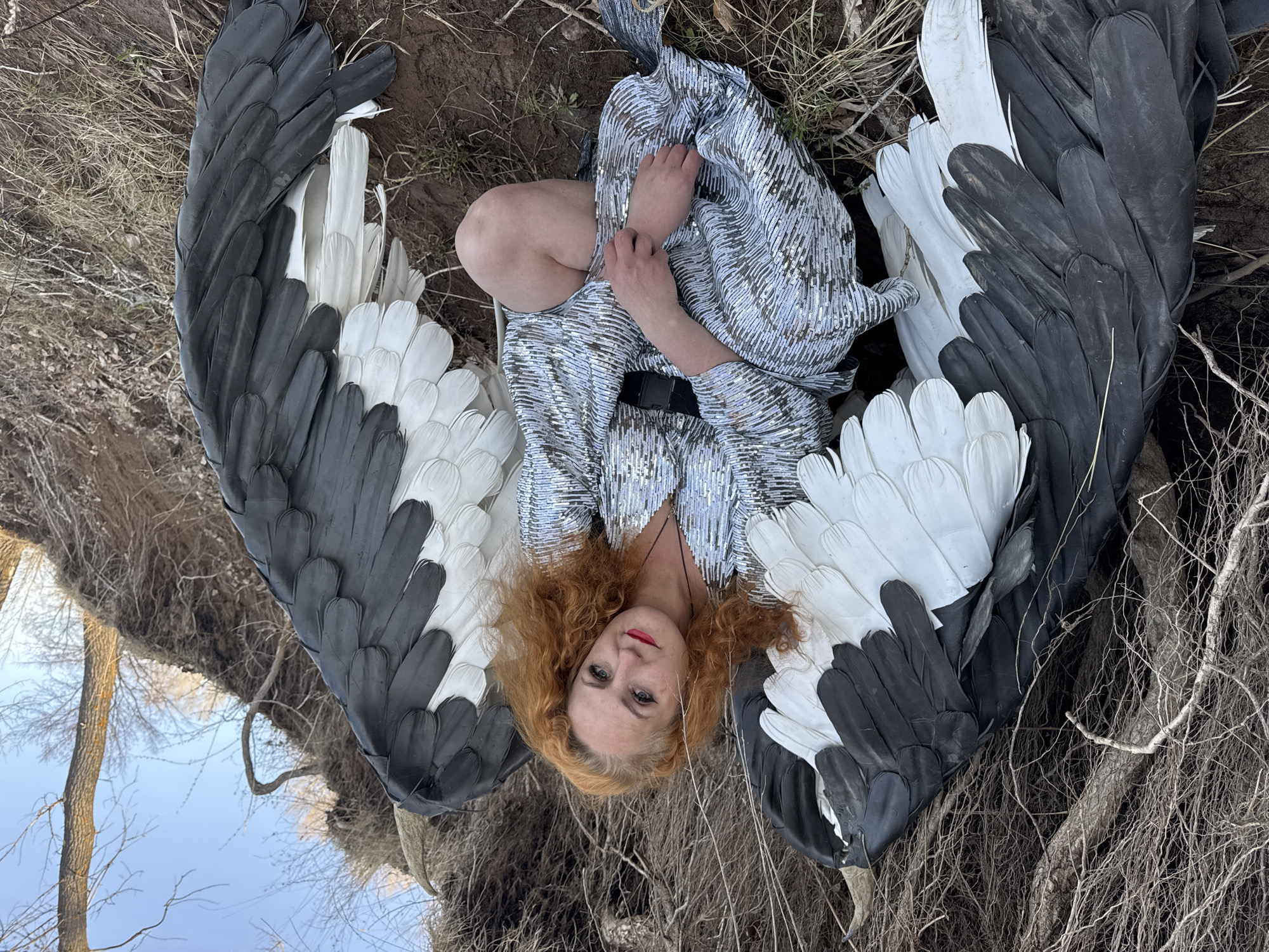 A woman lies on the ground, wearing a sequined dress and large black and white wings. Her red hair is loose, and she is holding her legs to her chest. The ground is covered in dry grass and dirt, with branches and roots visible. In the background, there is a glimpse of a tree and sky. The image appears to be taken outdoors, and based on the request, is likely located in Chaikovsky, Russia.