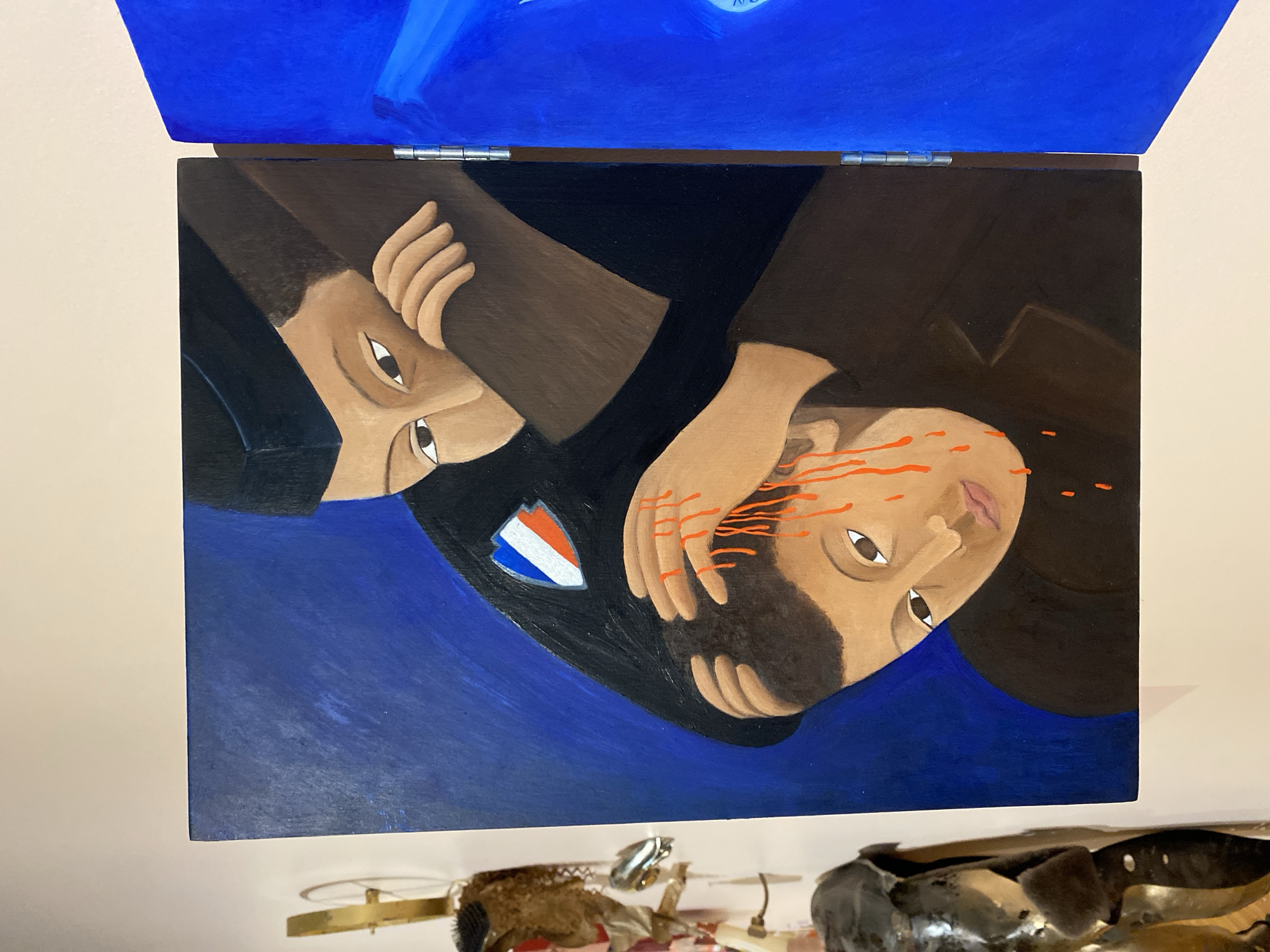 A vertically oriented painting is displayed against a light-colored wall, depicting two male figures with light brown skin tones against a solid deep blue background. The upper figure wears a dark uniform, a dark cap, and a shield-shaped patch on the left shoulder featuring red, white, and blue diagonal stripes. This figure's right arm is positioned around the lower figure's neck/shoulder area, and a hand from the upper figure is placed on the lower figure's head. Orange-red liquid, resembling blood, drips down from the lower figure's head and face. The lower figure wears a dark garment and has a somber expression. To the right of this painting, a second, entirely blue panel is visible, connected by silver metal hinges. To the left of the main painting, partially visible, are several objects: a golden, circular metal element at the top, and below it, various irregular, dark, and metallic or sculpted forms. The setting appears to be an indoor exhibition space or gallery. The precise location within Paris, France, cannot be determined from the provided image.