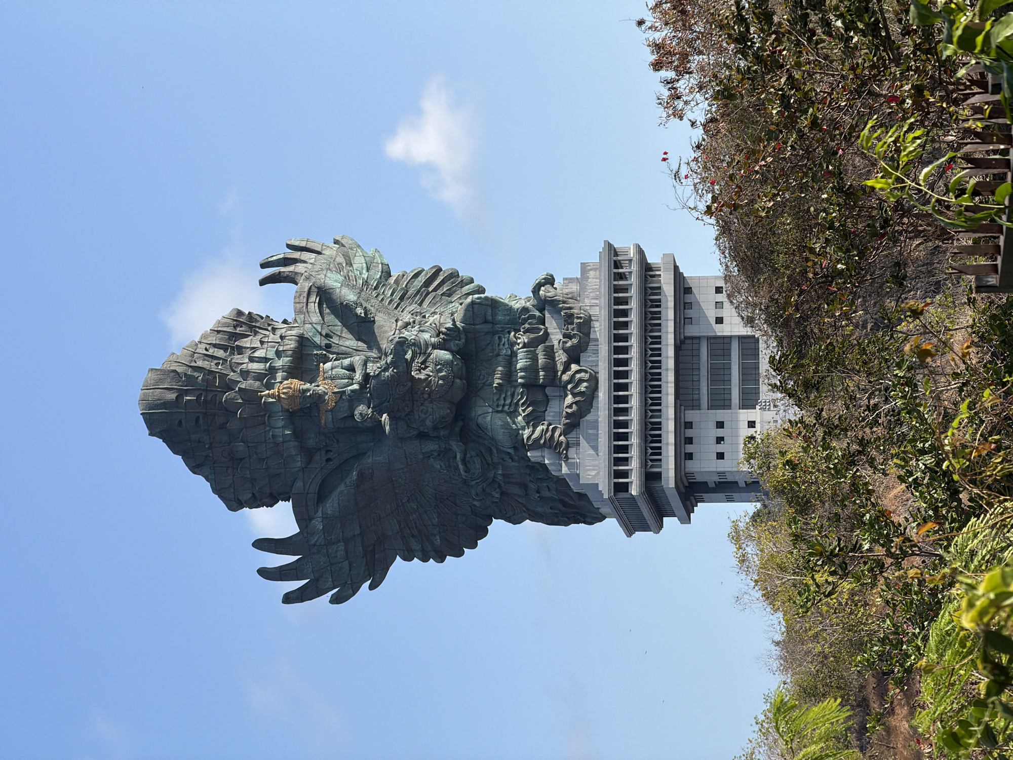 The image displays the colossal Garuda Wisnu Kencana statue in Ungasan, Kuta Selatan, Bali, Indonesia, set against a clear blue sky with scattered white clouds. The statue, predominantly bronze with a verdigris patina, depicts the Hindu deity Vishnu riding the mythical bird Garuda. It is perched atop a multi-story, modern building with a grey facade featuring a repetitive pattern of windows and decorative elements.
The lower portion of the frame is filled with dense, lush vegetation, including trees and shrubs with green and some reddish-brown leaves. A rustic, pointed wooden fence runs along the bottom edge of the image, partially obscuring the foreground foliage. The overall scene is one of grandiosity and natural beauty, with the towering statue dominating the landscape. There are no people visible in the image.
