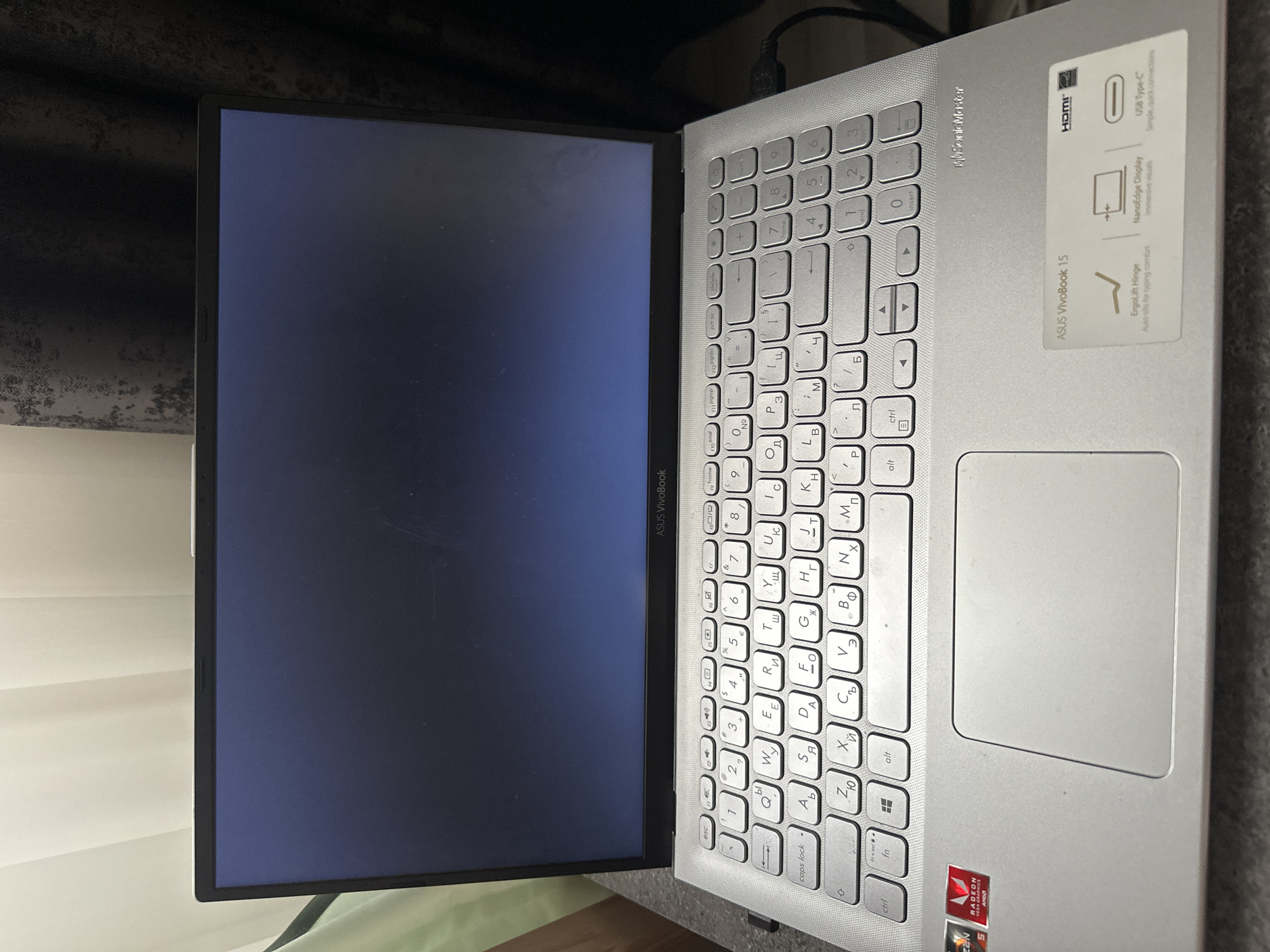 The image shows a silver ASUS VivoBook 15 laptop with a dark screen, positioned on a grey textured surface, likely a desk. The laptop is open, with the keyboard visible. The keyboard features Cyrillic characters alongside Latin letters, indicating it is likely a Russian or Ukrainian layout. Stickers on the laptop indicate it has Radeon Vega Graphics and SonicMaster audio. The background consists of a light-colored curtain on the left and a dark, textured curtain on the right, suggesting the laptop is indoors. There are no people or animals visible in the frame. The lighting is dim, with a slight glare on the laptop screen.