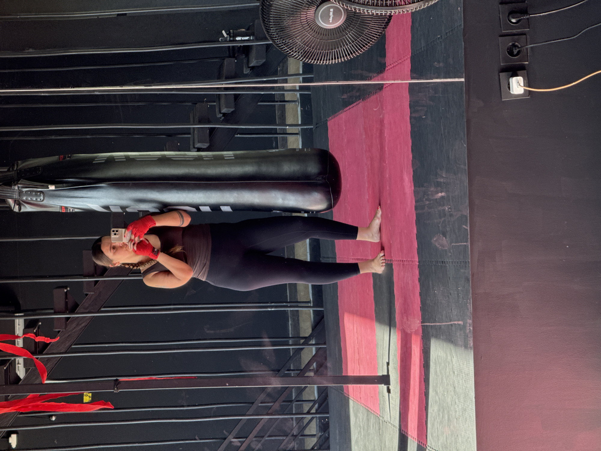 In a gym in Ubud, Indonesia, a woman wearing a dark tank top, black leggings, and red boxing gloves stands in front of a large black punching bag, taking a selfie with her phone. The gym features a black metal staircase with red ribbons hanging from it, a black fan, and red and black interlocking mats on the floor. The scene is illuminated by sunlight casting shadows on the floor. Electrical outlets are visible on a dark wall in the lower right corner of the frame.