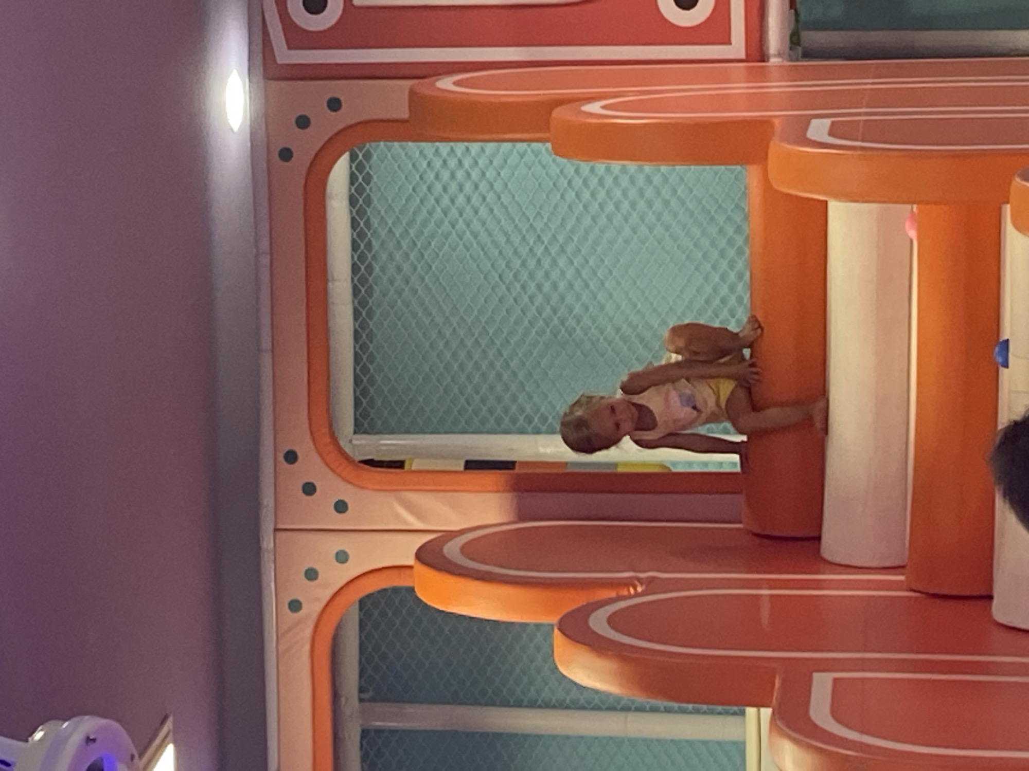 The image shows a young child sitting on the edge of a colorful play structure. The child, appearing to be a girl with blonde hair, is wearing a tank top and shorts. She is positioned on a padded orange step, looking down and slightly to her right. The play structure itself is bright orange with white trim and features curved elements and steps. Behind the child is a turquoise netted wall. The ceiling is a light purple color, and there are lights visible in the upper portion of the frame. The overall environment appears to be an indoor playground. The location is described as Phu Thuy, Vietnam.