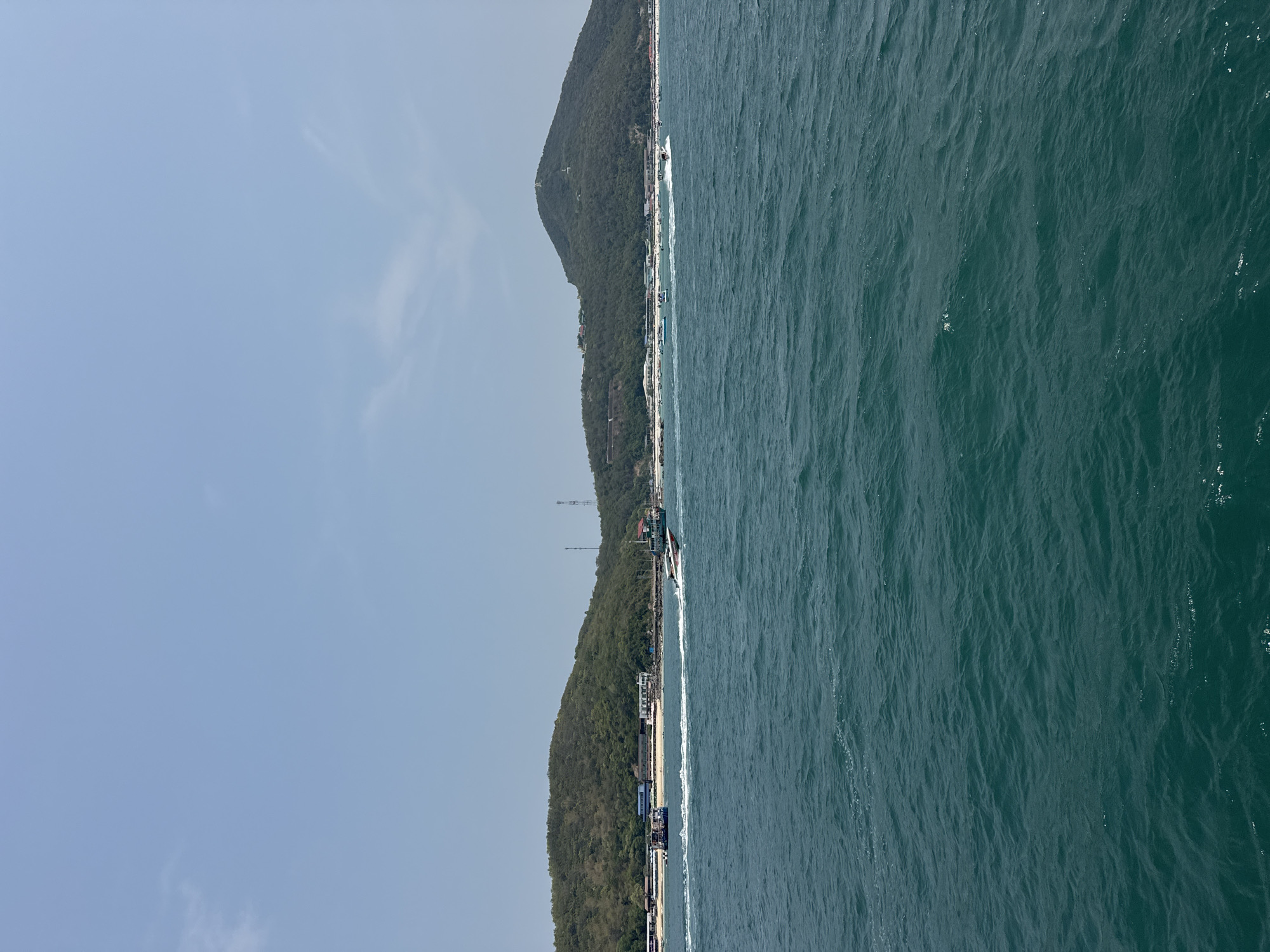 The image shows the coastline of Pattaya City, Thailand, with a densely vegetated mountain range running parallel to the shore. The sea occupies the right half of the image, exhibiting a dark, rippled surface. A narrow strip of beach is visible along the coastline, lined with buildings. A boat is travelling along the coastline. The sky is a pale blue with few clouds.