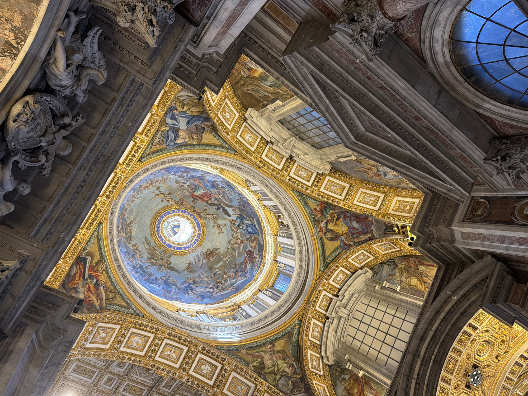 The image shows an upward view of the interior of St. Peter's Basilica in Vatican City. The ornate ceiling features a large dome with a circular opening at its apex, allowing light to enter. The dome is adorned with detailed frescoes depicting religious figures and scenes, painted in blue, gold, and beige tones. Around the dome are layers of intricate architectural details, including gilded squares and arched windows. Statues and carvings are present on the walls and columns, adding to the basilica's opulent design. The overall color palette is dominated by golds, browns, and blues.