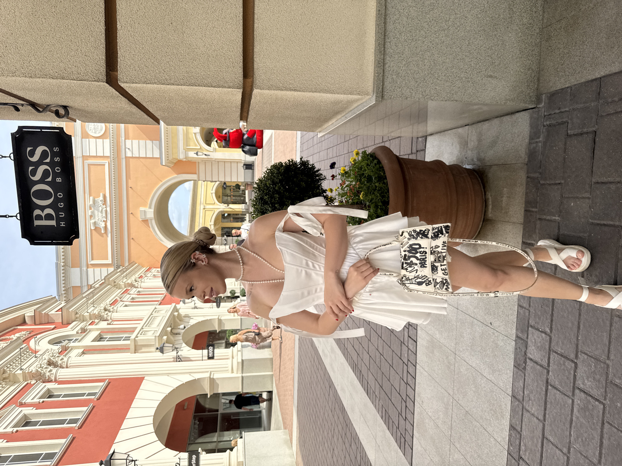 A blonde woman, facing left in profile, stands on a paved walkway. She is wearing a white off-the-shoulder dress with tie details on the sleeves, white pearl necklaces, and white sandals. She is holding a white handbag with black graffiti-style lettering. Behind her, an outdoor shopping area features buildings with arched entrances and decorative architectural details. A large potted plant with dark green foliage and colorful flowers sits to her right. A black sign with "BOSS HUGO BOSS" in white letters hangs above the scene. The ground is paved with alternating patterns of dark and light gray bricks. The woman's expression is neutral as she looks towards the left. The context of Saint Petersburg, Russia, is not discernible from the visual elements of the image.