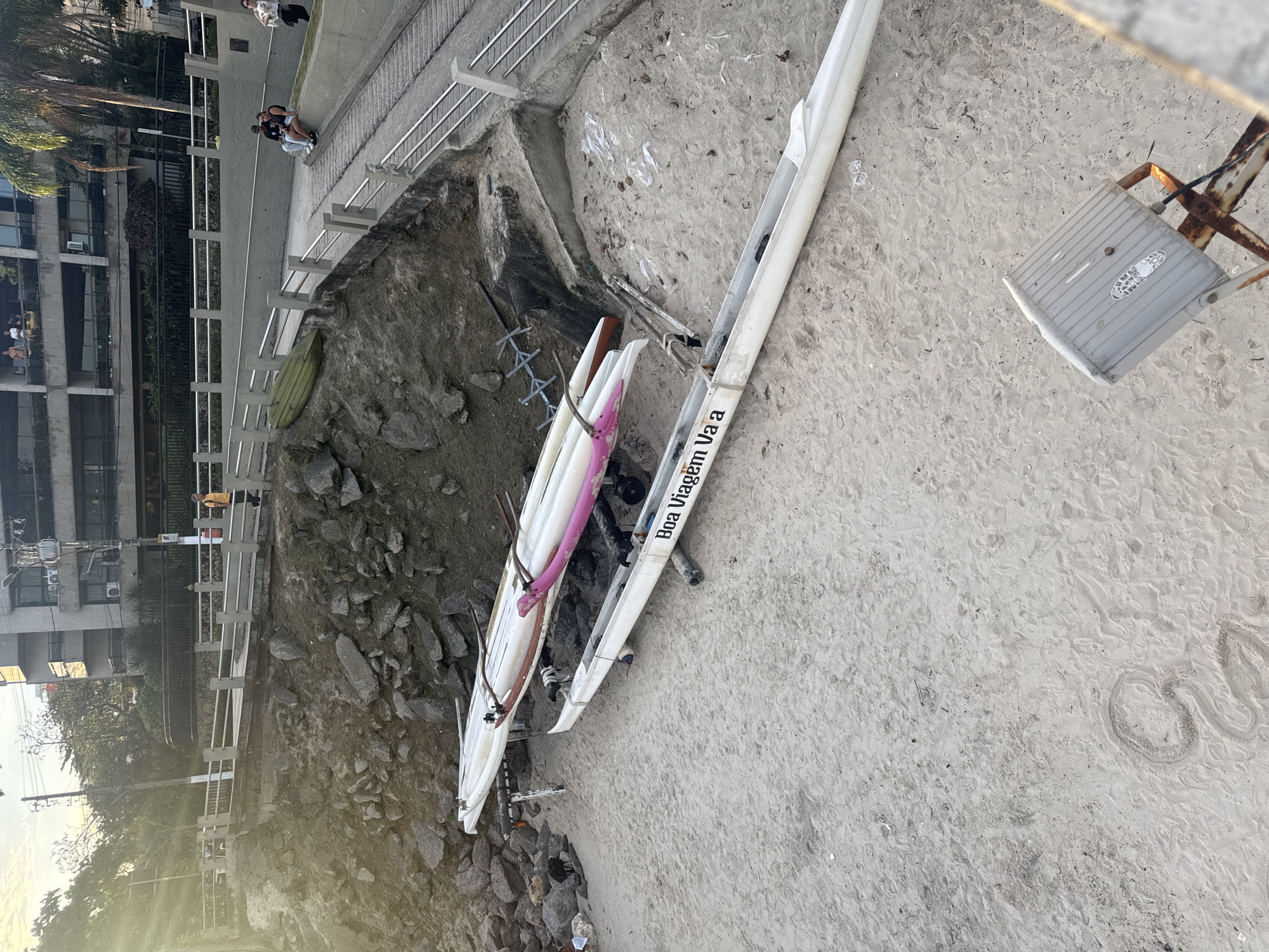 The image shows two canoes on a sandy beach in Niterói, Brazil. One canoe is white with pink accents and is positioned on a trailer. The other canoe is white and empty, placed on the sand. In the background, there is a promenade with a railing and a slope leading up to it. Several people are visible on the promenade, some sitting and others standing. A building with balconies is seen further in the background. The environment appears to be a coastal area with some rocky terrain and vegetation. The lighting suggests it is either early morning or late afternoon. A grey metal fixture with a lamp is also present on the sand.