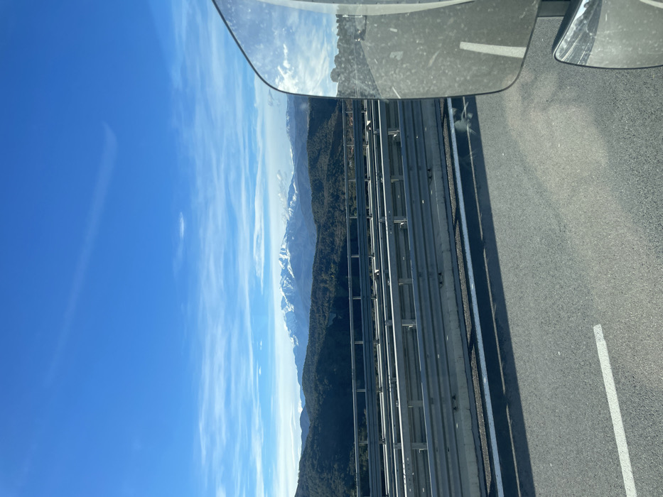 A view from a high bridge shows a road with a guardrail and a distant mountain range. The peaks are dusted with snow against a blue sky. A rearview mirror is visible in the upper left corner, reflecting the sky. The road surface is gray, marked with white lines, suggesting a highway. The scene implies travel, with nature's grandeur as a backdrop.
