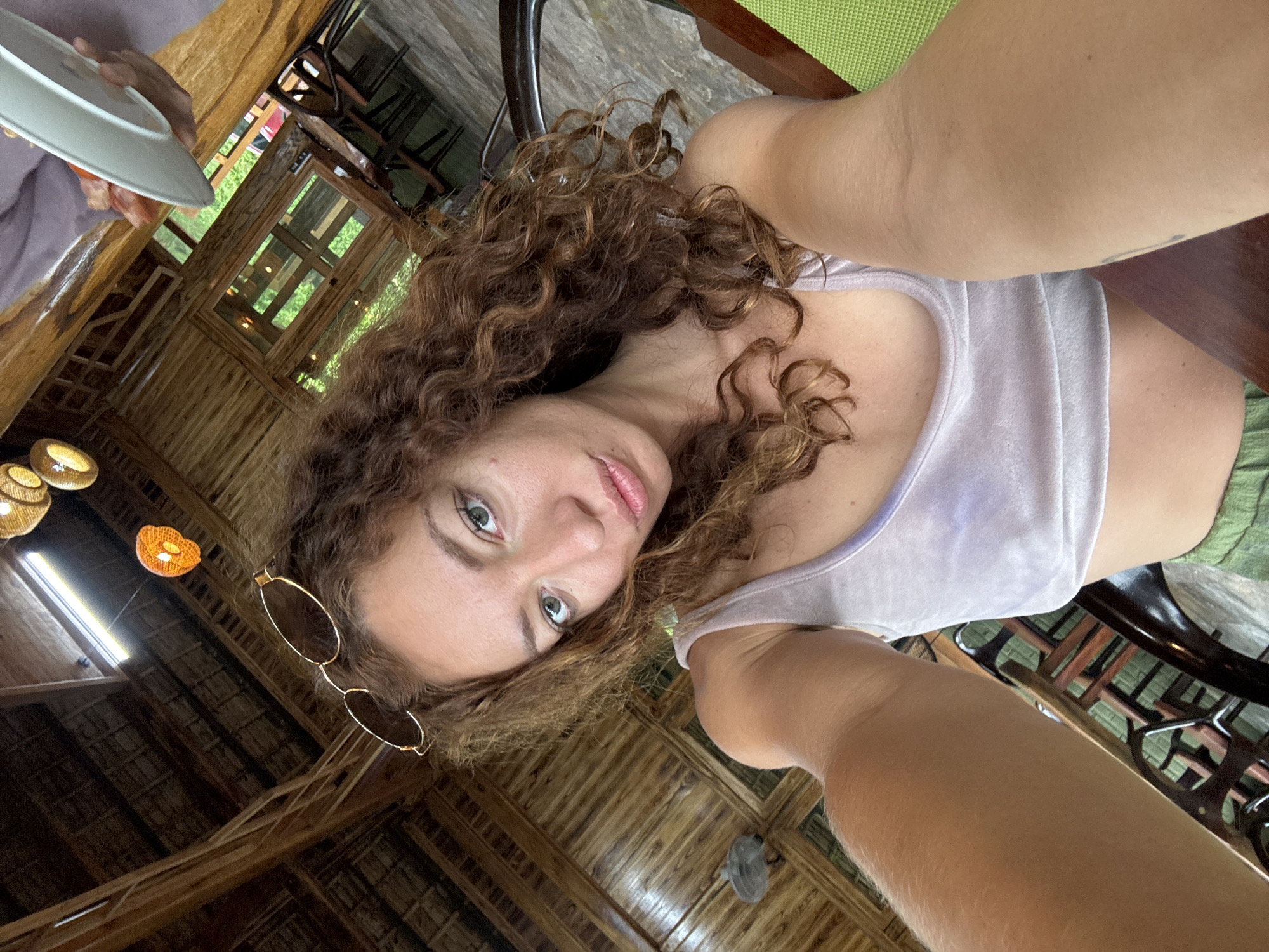 The image shows a woman taking a selfie in what appears to be an open-air restaurant in Pù Luông Commune, Vietnam. The woman has curly brown hair and is wearing a light purple crop top and green pants. She has sunglasses on her head. The background is a wooden structure with a thatched roof, and tables and chairs are visible. There are also hanging light fixtures and a small fan mounted on the wall. The scene suggests a tropical or subtropical environment.