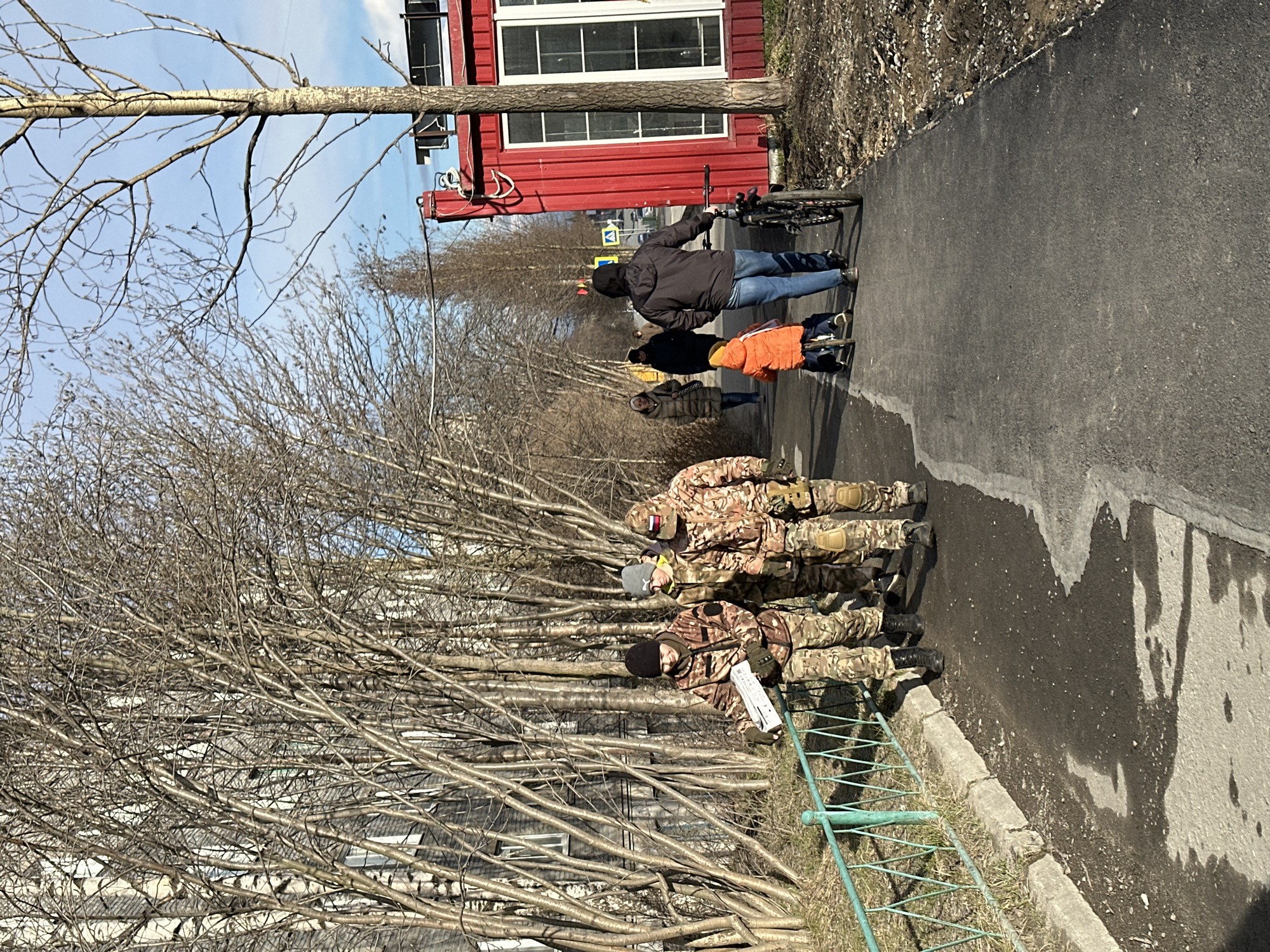 The image shows a group of people walking on a paved road in what appears to be a residential area. Three individuals dressed in camouflage military uniforms stand near a green metal fence to the left of the road. One of them is holding a white object that appears to be paper or a document. Ahead of them, a group of civilians, including a person in an orange jacket and another pushing a bicycle, are walking in the same direction. A red building with white-framed windows is visible on the right side of the road. Bare tree branches fill the left side of the image, partially obscuring the view of buildings in the background. The weather seems clear, with a blue sky visible in the upper part of the image. The location is described as Unknown, Unknown.
