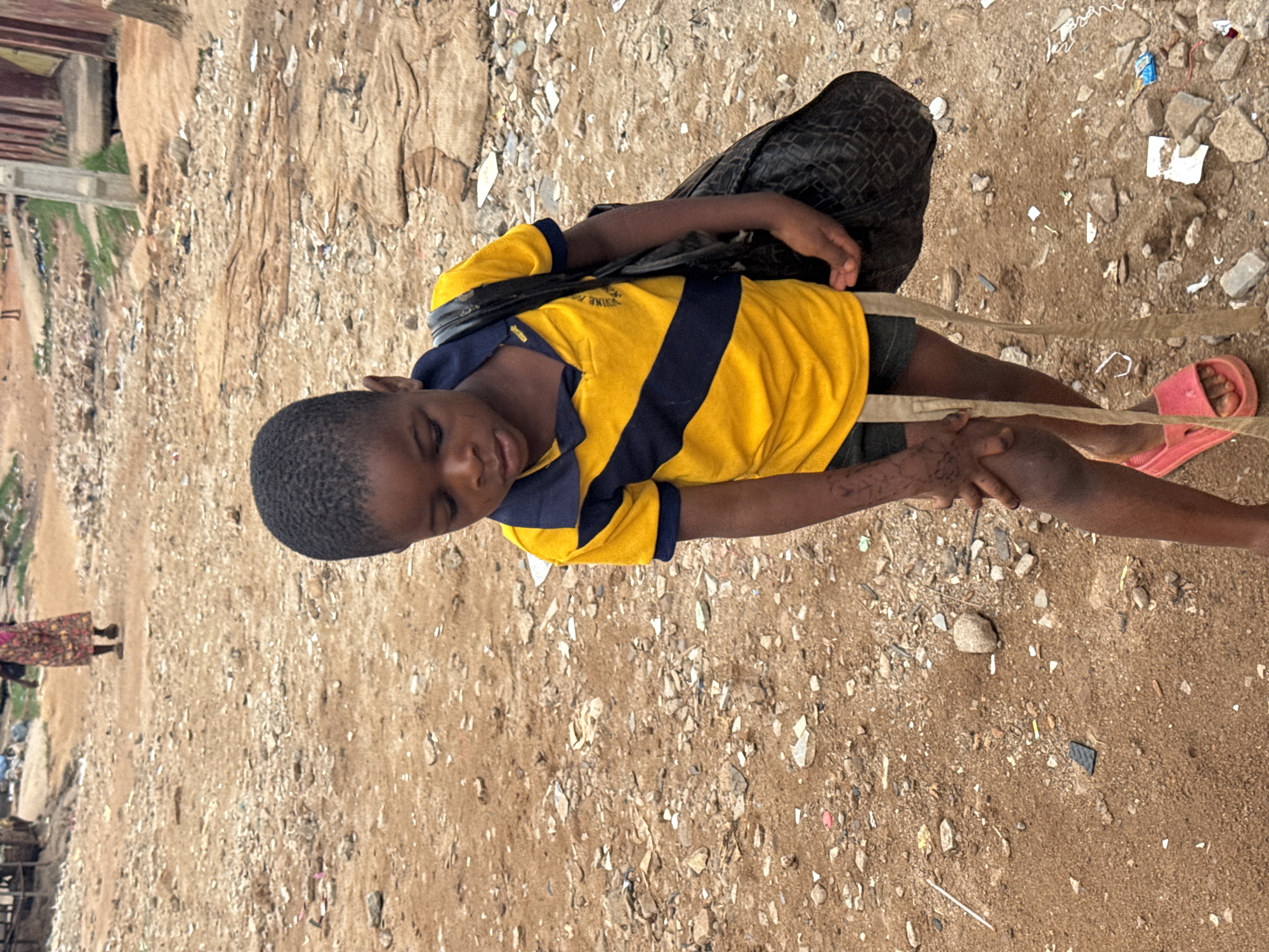 In Onitsha, Nigeria, a child in a yellow and blue striped shirt, black shorts, and pink sandals stands on a gravel and dirt ground, holding a light brown cord. The child wears a black bag. The ground is littered with debris. In the background are buildings, greenery, and a woman in a patterned dress.