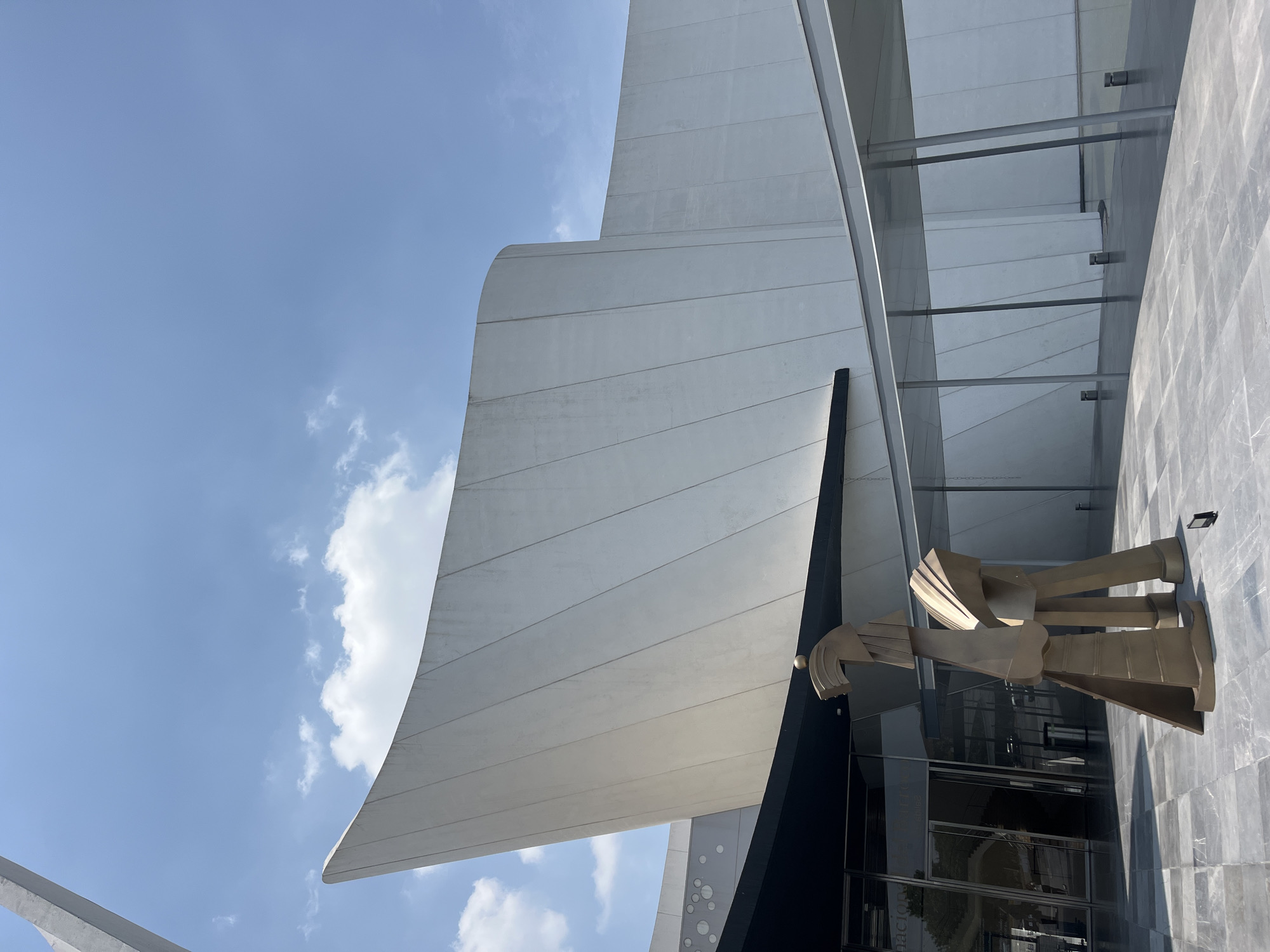 The image shows an architectural view of the Museo Internacional Barroco (International Baroque Museum) in San Bernardino Tlaxcalancingo, Mexico. The composition is angled upwards, capturing a portion of the building's exterior against a blue sky with scattered clouds. Key features include a large, white, curved structural element, likely part of the building's facade, and a contemporary metal sculpture consisting of several abstracted shapes. The building also features glass walls, partially visible.
