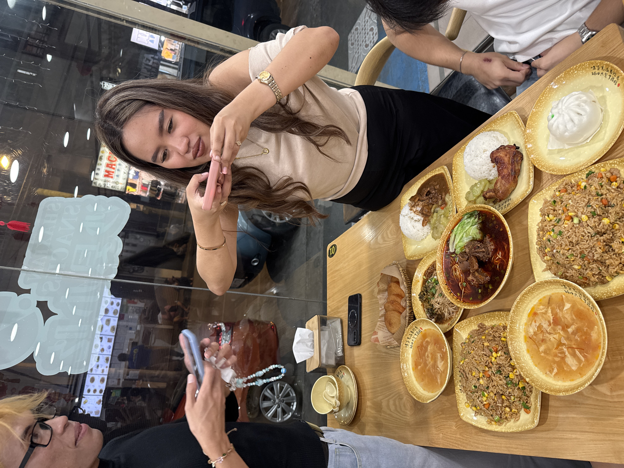 The image depicts a scene inside a restaurant in Davao City, Philippines. Three people are visible: a woman with long brown hair holding a pink object (possibly a phone or lipstick), a person with short blonde hair holding a blue phone, and another person partially visible at the top right of the frame.
A wooden table is covered with several plates of food, including rice, fried rice, a steamed bun, a dish with meat and vegetables in a sauce, and soup. Other objects on the table include a box of tissues, a phone, a cup and saucer, and a small basket containing bread.
The woman with brown hair is the main focus, captured mid-action as if inspecting the pink object in her hands. The person with blonde hair is holding a phone, perhaps taking a photo or video. The third person appears to be seated and looking down at the table.
The background includes a window displaying a menu board and reflections of the street scene outside. The restaurant's lighting creates highlights on the glass and tableware.